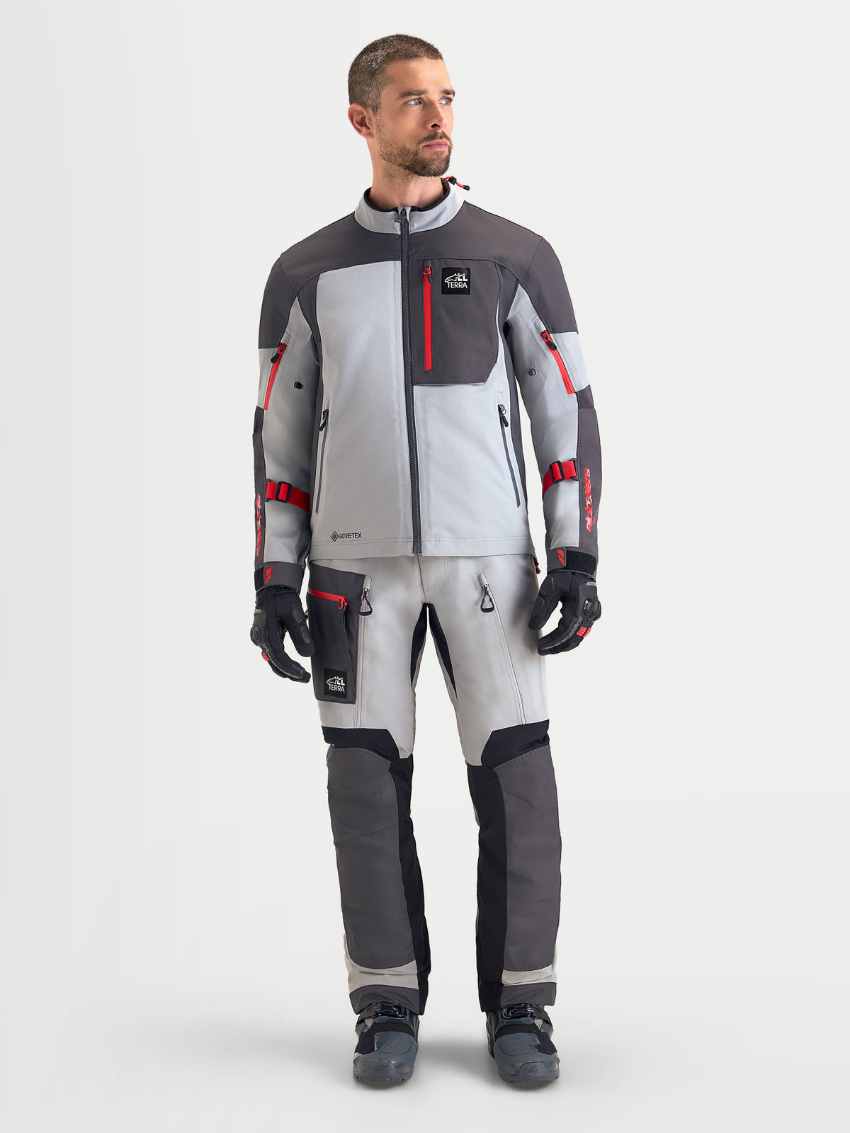 Alpinestars Arenis Gore-Tex jacka, äventyrsmotorcykeljacka för herr i mellergrå, mörkgrå och svart, framifrån på modell som visar vattentät textilkonstruktion och röda dragkedjedetaljer