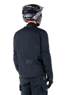 Alpinestars St-7 Prime GORE-TEX-jacka, svart, vy bakifrån på en förare som bär jackan med matchande hjälm och byxor, med Alpinestars-logotyp och geografiska koordinater på nedre ryggen.