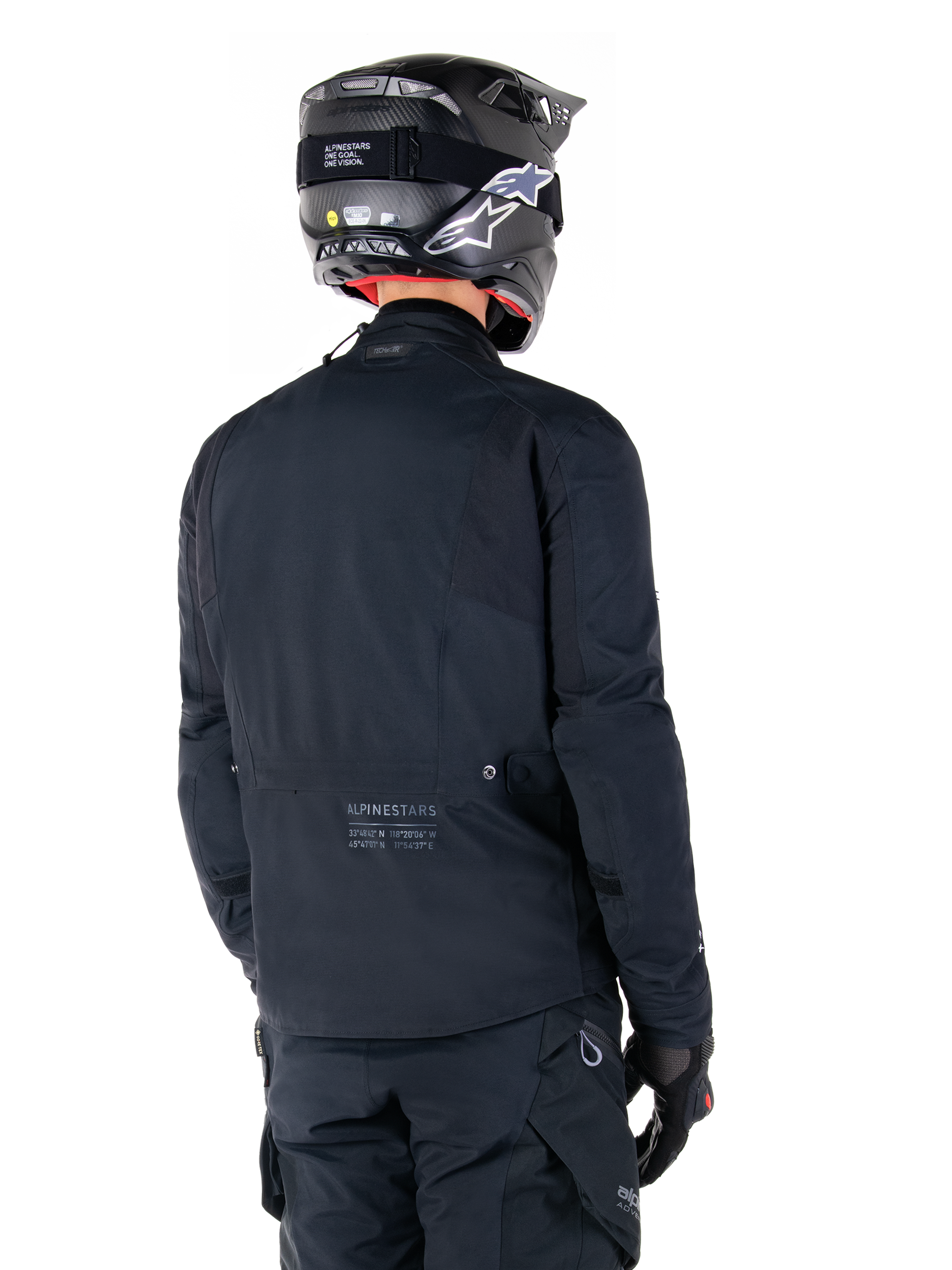 Alpinestars St-7 Prime GORE-TEX-jacka, svart, vy bakifrån på en förare som bär jackan med matchande hjälm och byxor, med Alpinestars-logotyp och geografiska koordinater på nedre ryggen.