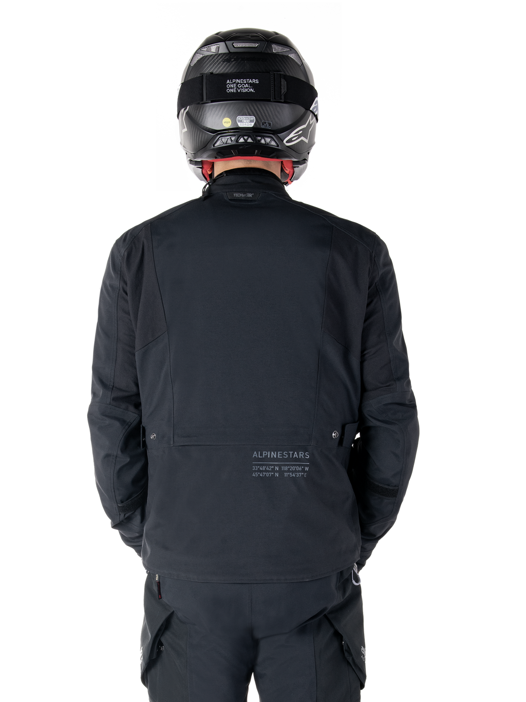 Alpinestars St-7 Prime GORE-TEX-jacka, svart, vy bakifrån av en förare med subtil logotyp och geografiska koordinater på nedre delen av ryggen, bärs med matchande svart hjälm och byxor, stilren och teknisk motorcykelutrustning