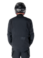 Alpinestars St-7 Prime GORE-TEX-jacka, svart, vy bakifrån av en förare med subtil logotyp och geografiska koordinater på nedre delen av ryggen, bärs med matchande svart hjälm och byxor, stilren och teknisk motorcykelutrustning