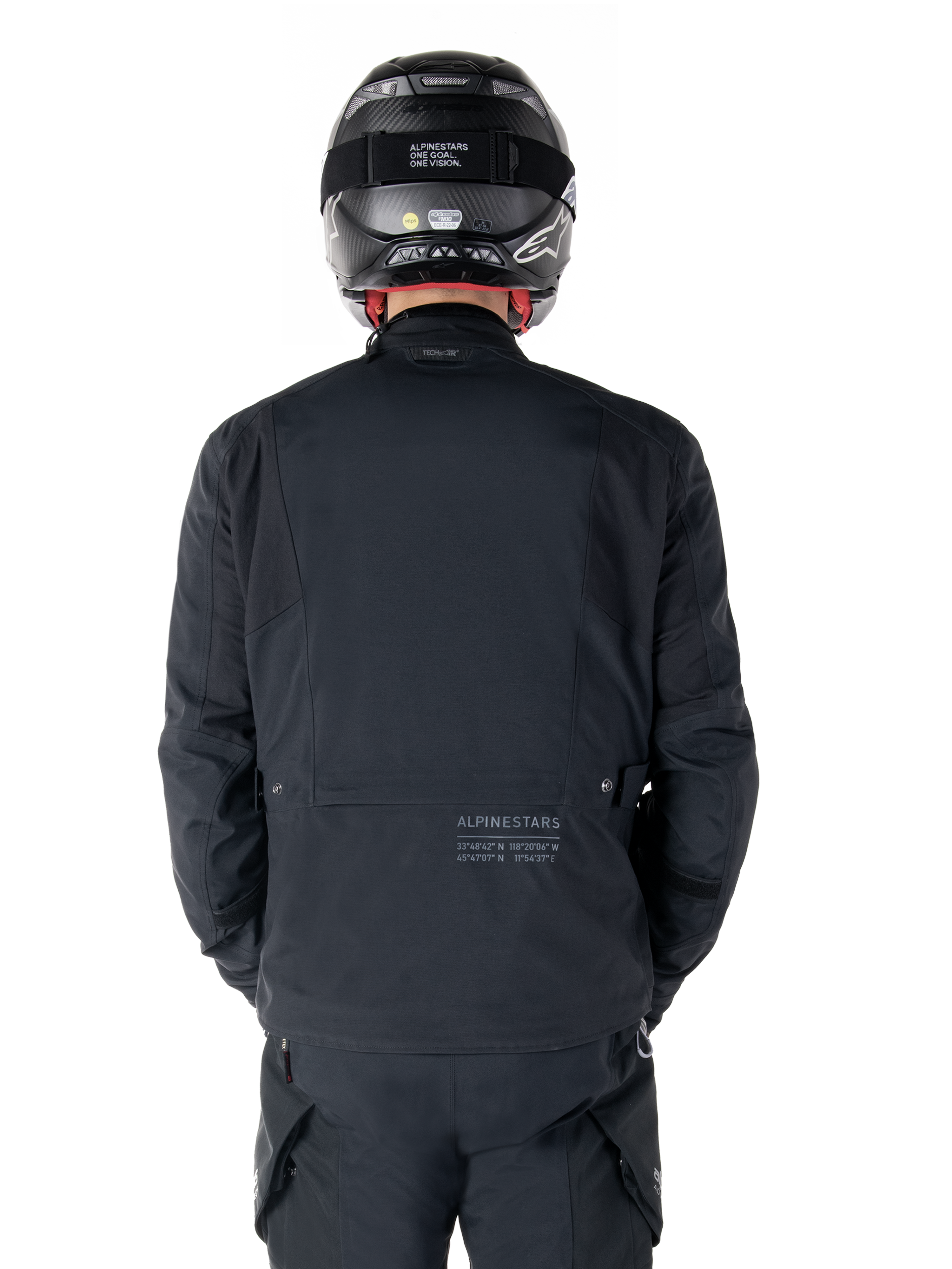 Alpinestars St-7 Prime GORE-TEX-jacka, svart, vy bakifrån av en förare med subtil logotyp och geografiska koordinater på nedre delen av ryggen, bärs med matchande svart hjälm och byxor, stilren och teknisk motorcykelutrustning