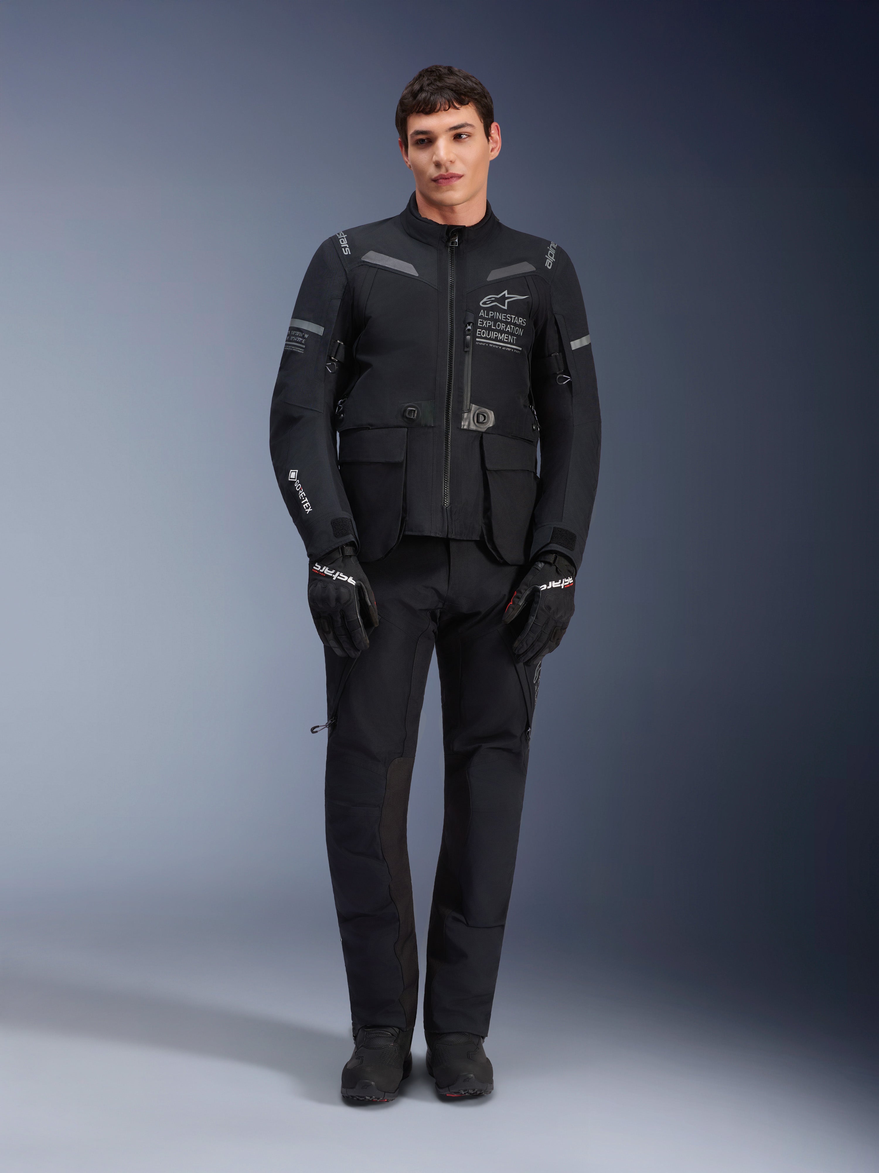 Alpinestars ST-7 2L GORE-TEX-jacka, motorcykeljacka för adventure-touring, svart och mörkgrå, bärs med matchande svarta körbyxor och handskar, med flera cargo-fickor, reflexdetaljer och Alpinestars Exploration Equipment-logotyp på bröstet, funktionell design för väg- och terrängkörning
