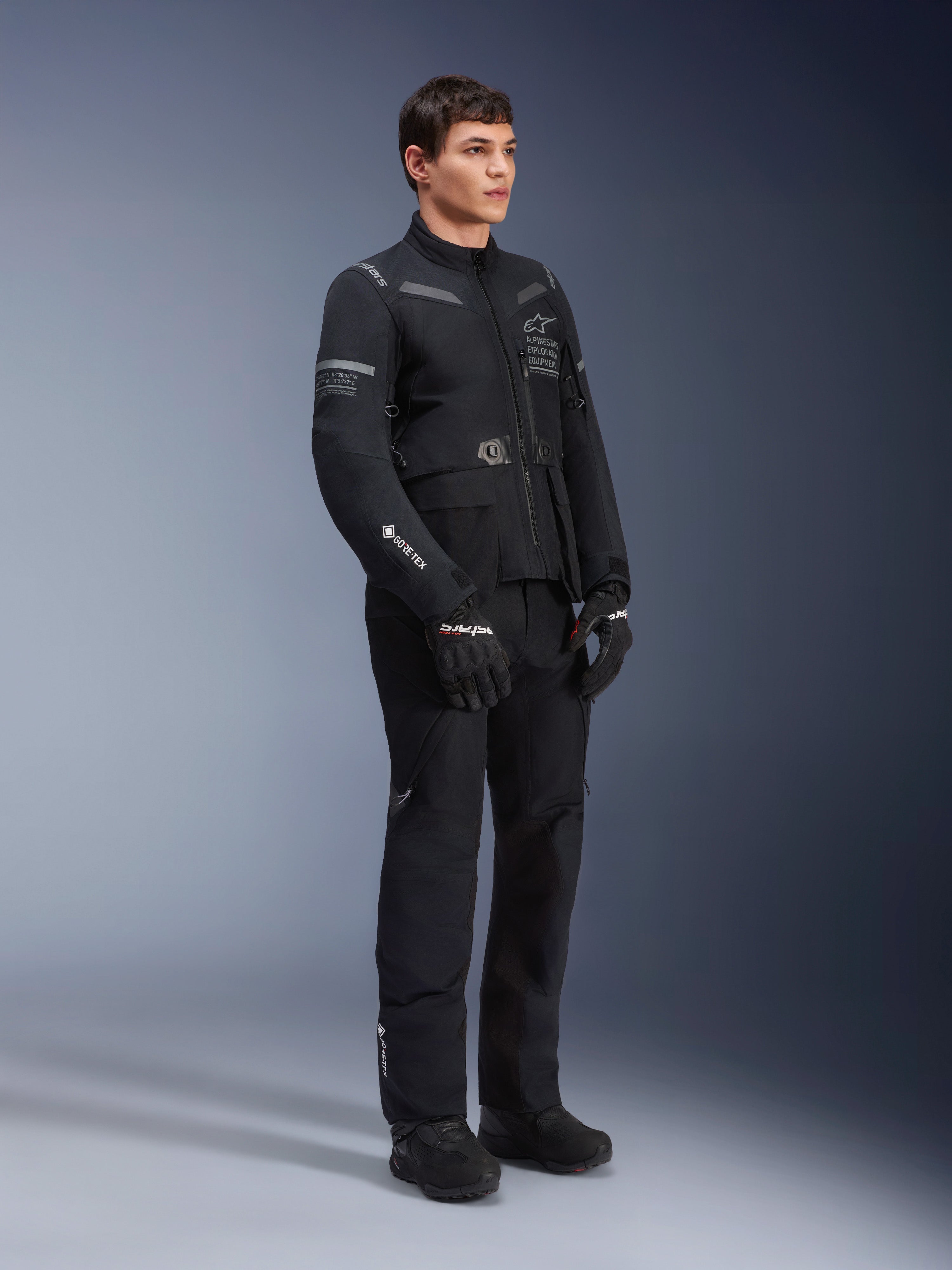 Manlig modell iförd Alpinestars ST-7 2L GORE-TEX-jacka i Black Dark Gray, motorcykeljacka för äventyr och touring, matchande svarta byxor och handskar, trekvarts helkroppsvy, med GORE-TEX-logotyp och Alpinestars Exploration Equipment-märkning