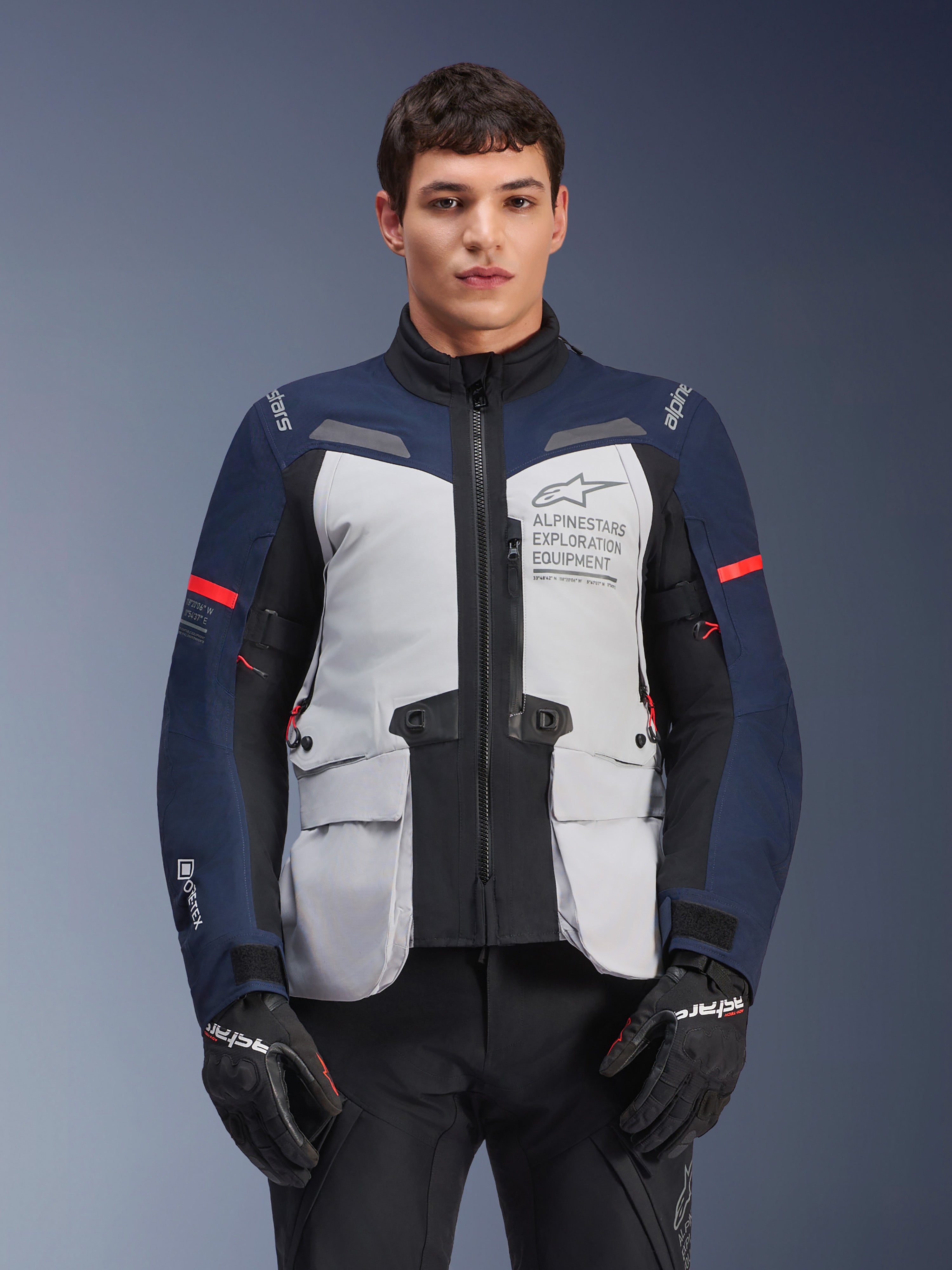 Alpinestars ST-7 2L GORE-TEX-jacka, motorcykeljacka för äventyrstouring, isgrå och mörkblå med röda detaljer, vattentät och andningsbar, bärs av en modell med svarta handskar, med Alpinestars Exploration Equipment-logotyp på bröstet
