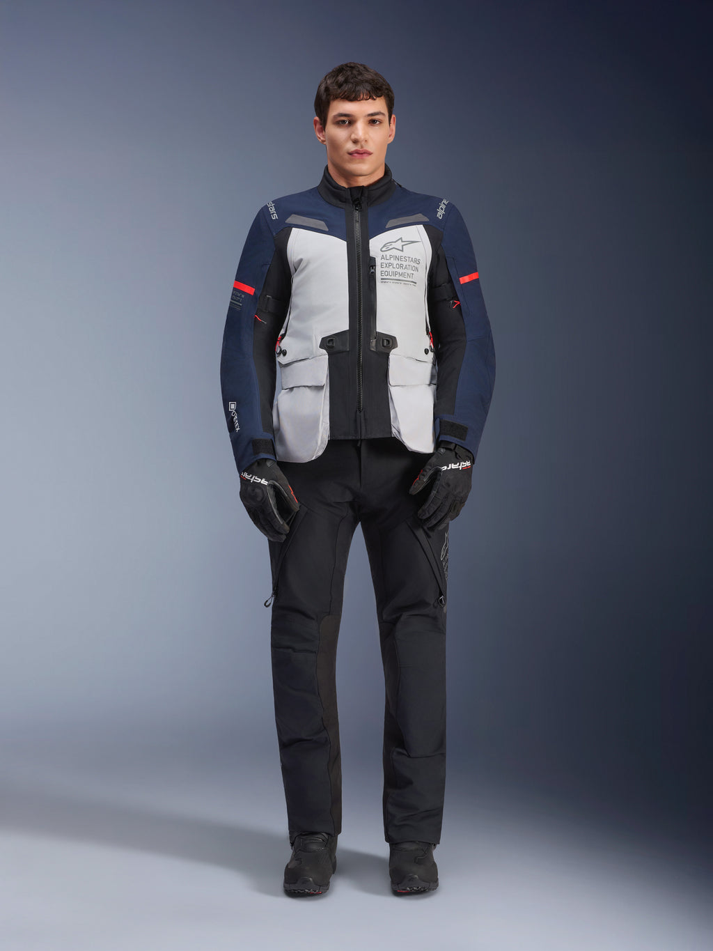 Alpinestars ST-7 2L GORE-TEX-jacka, motorcykeljacka för adventure touring, isgrå, mörkblå och svart, bärs med svarta byxor och handskar, stora utanpåliggande fickor fram, röda detaljer på ärmarna, laminerat GORE-TEX-skal för vattentät prestanda.