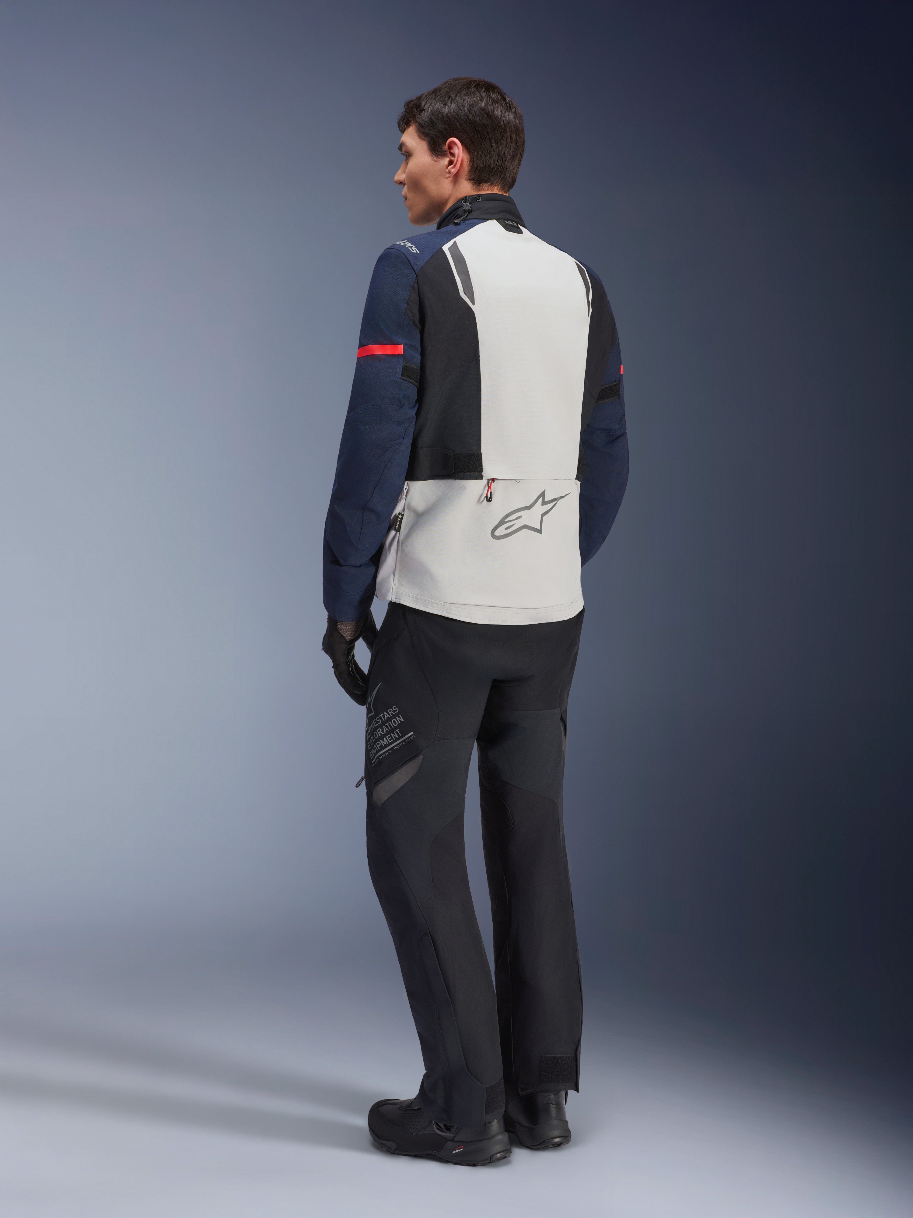 Alpinestars ST-7 2L GORE-TEX-jacka i Ice Gray, Dark Blue och Black, vy bakifrån på modell, ljusgrå ryggpanel med Alpinestars-logotyp, mörkblå ärmar med röd rand, kombinerad med svarta adventure-byxor och stövlar, motorcykel-touringutrustning