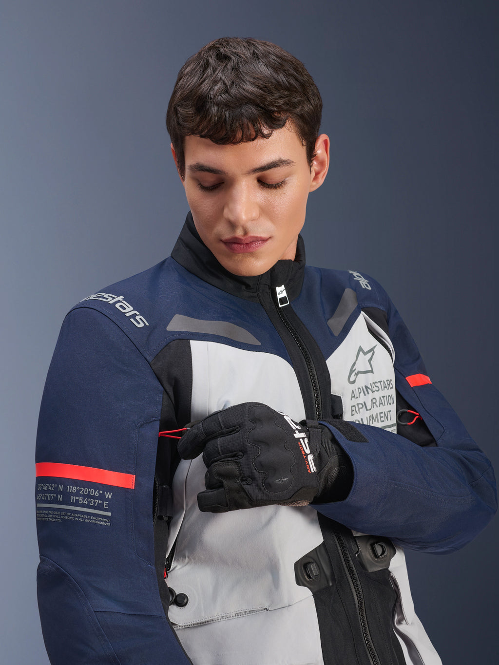 Alpinestars ST-7 2L GORE-TEX-jacka, isgrå, mörkblå och svart med orangea detaljer, bärs av en man som justerar en detalj på ärmen, med 'Alpinestars Exploration Equipment'-logotyp och geografiska koordinater, professionell MC-utrustning för äventyrskörning