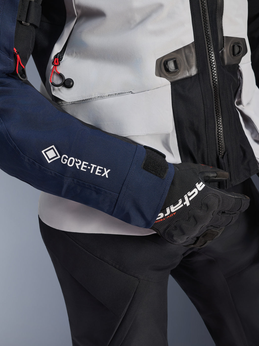 Alpinestars ST-7 2L GORE-TEX-jacka, närbild på mörkblå ärm med vit GORE-TEX-logotyp, isgrå torso med svarta detaljer och röda dragskodetaljer, bärs med svarta Alpinestars-handskar och byxor, adventure touring-motorcykelutrustning