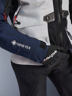 Alpinestars ST-7 2L GORE-TEX-jacka, närbild på mörkblå ärm med vit GORE-TEX-logotyp, isgrå torso med svarta detaljer och röda dragskodetaljer, bärs med svarta Alpinestars-handskar och byxor, adventure touring-motorcykelutrustning