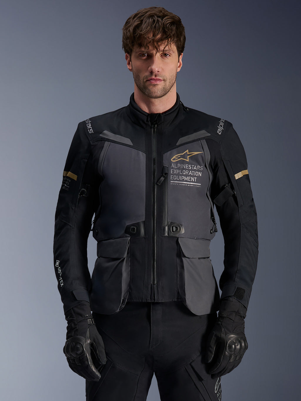 Alpinestars ST-7 2L GORE-TEX-jacka, äventyrstouring-motorcykeljacka för herrar, mörkgrå och svart med khaki-detaljer, med "Alpinestars Exploration Equipment"-branding, vattentäta cargo-fickor och hög krage, bärs av en modell med svarta handskar
