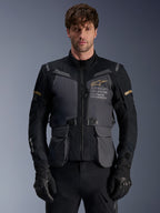 Alpinestars ST-7 2L GORE-TEX-jacka, äventyrstouring-motorcykeljacka för herrar, mörkgrå och svart med khaki-detaljer, med "Alpinestars Exploration Equipment"-branding, vattentäta cargo-fickor och hög krage, bärs av en modell med svarta handskar