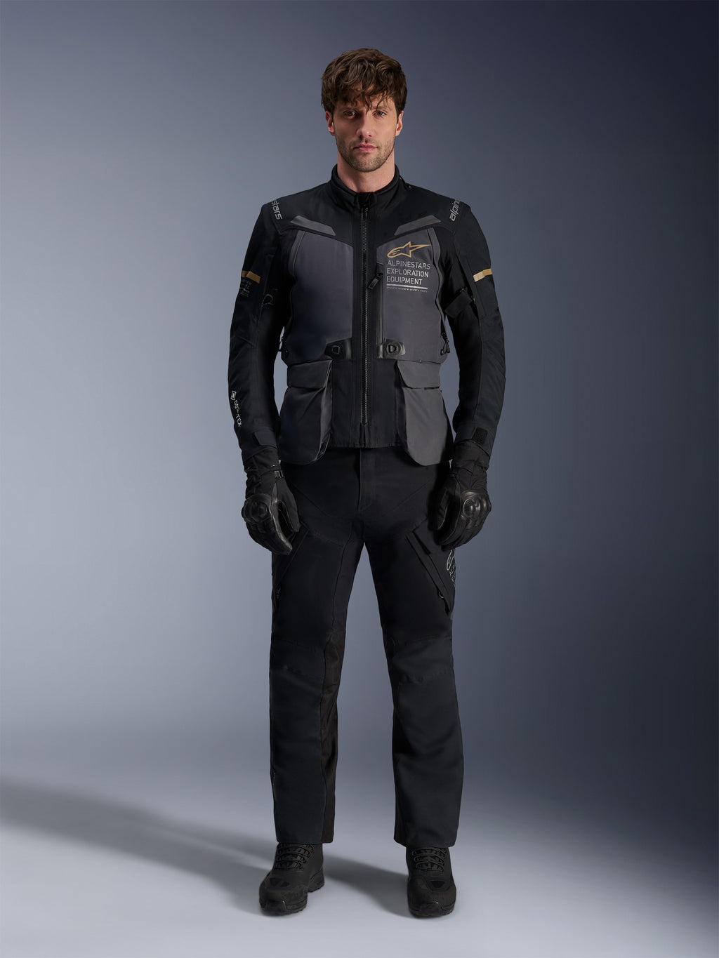 Manlig modell iklädd Alpinestars ST-7 2L GORE-TEX-jacka i färgen Deep Gray Black Dark Khaki, med 'ALPINESTARS EXPLORATION EQUIPMENT'-logotyp på bröstet, matchad med svarta mc-byxor, handskar och stövlar, studiomiljö, vy framifrån.