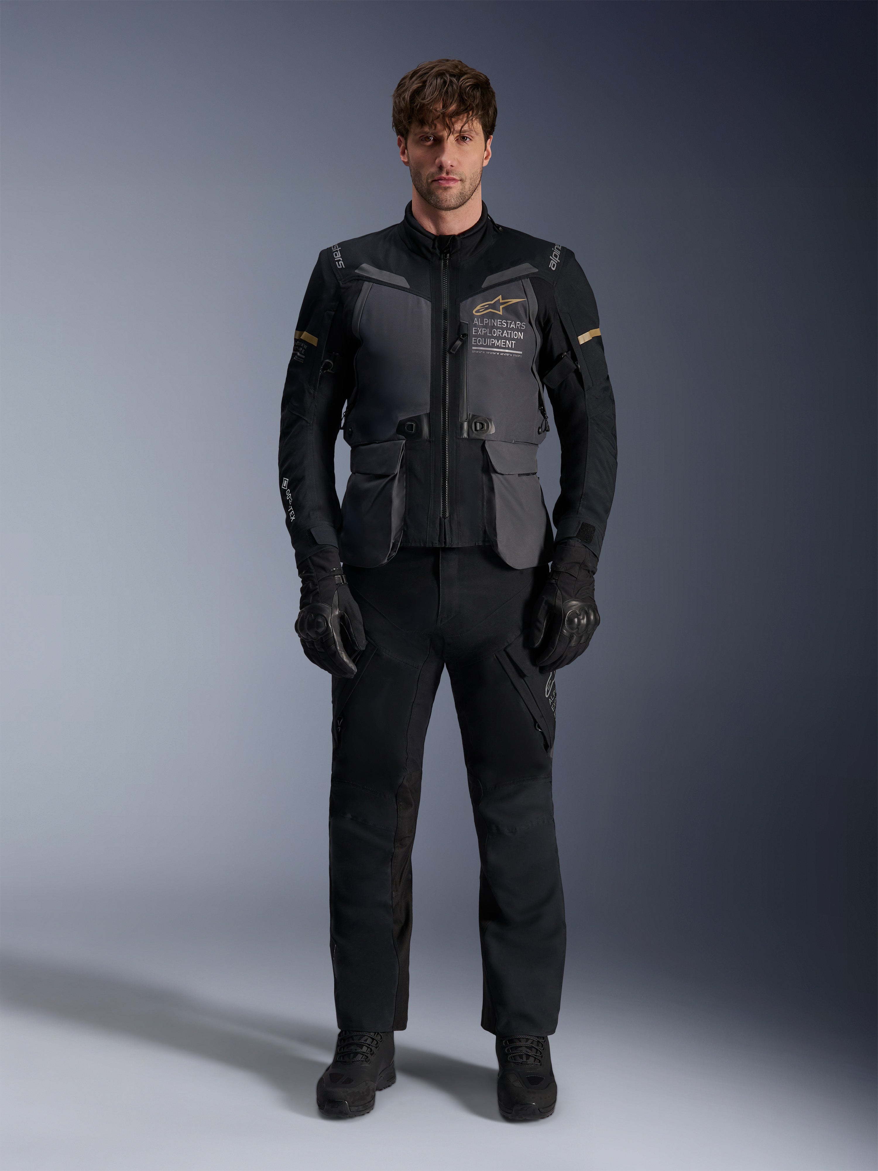 Manlig modell iklädd Alpinestars ST-7 2L GORE-TEX-jacka i färgen Deep Gray Black Dark Khaki, med 'ALPINESTARS EXPLORATION EQUIPMENT'-logotyp på bröstet, matchad med svarta mc-byxor, handskar och stövlar, studiomiljö, vy framifrån.