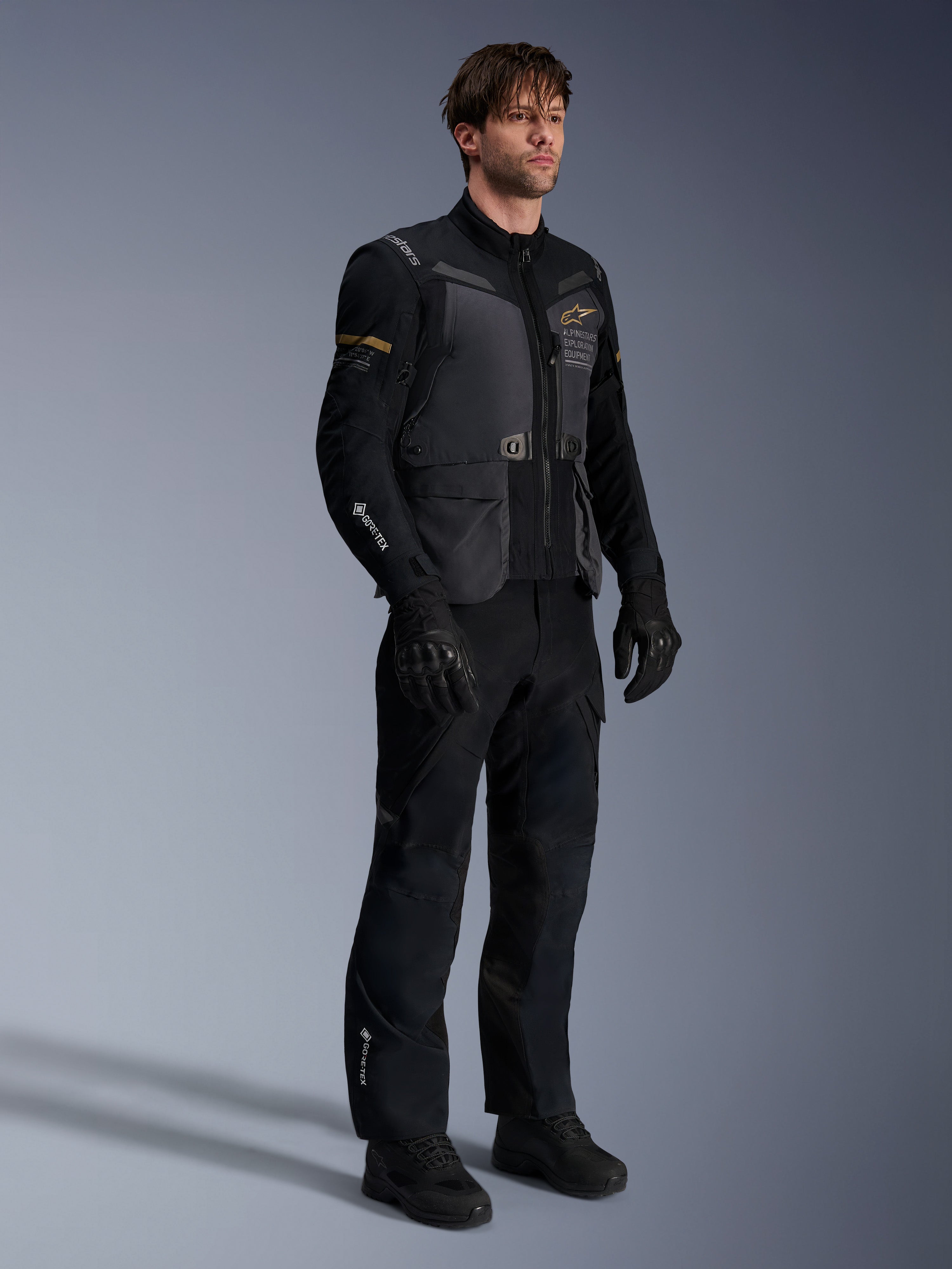 Manlig modell som bär Alpinestars ST-7 2L GORE-TEX-jacka i Deep Gray Black och Dark Khaki, kombinerad med svarta adventure-byxor och handskar, vattentätt laminerat skal med cargo-fickor och teknisk design, idealisk för adventure touring.