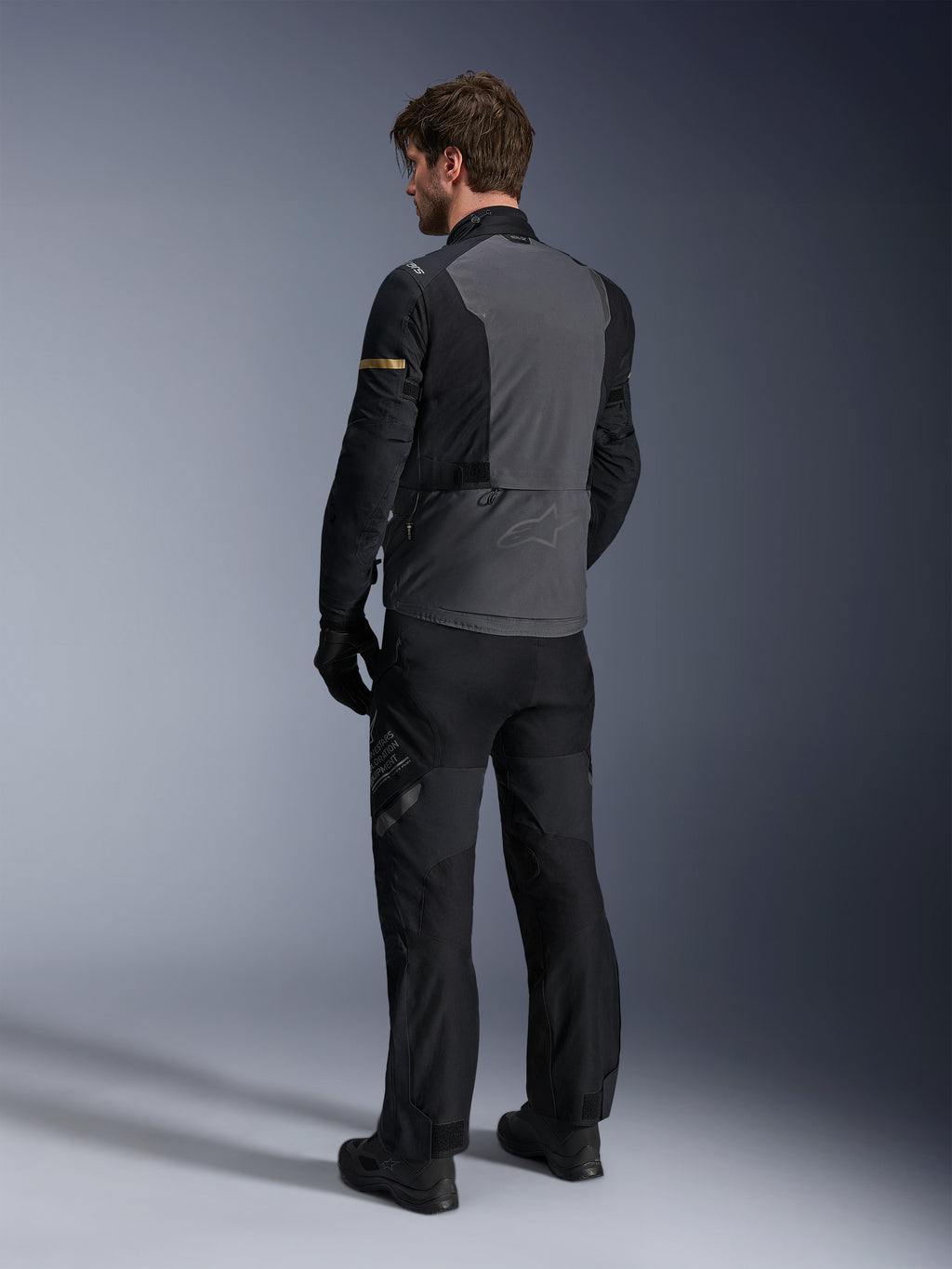 Alpinestars ST-7 2L GORE-TEX-jacka, mörkgrå och svart med mörka kaki-detaljer, vy bakifrån på en förare som bär jackan och matchande motorcykelbyxor, adventure touring-utrustning med vattentät laminerad konstruktion