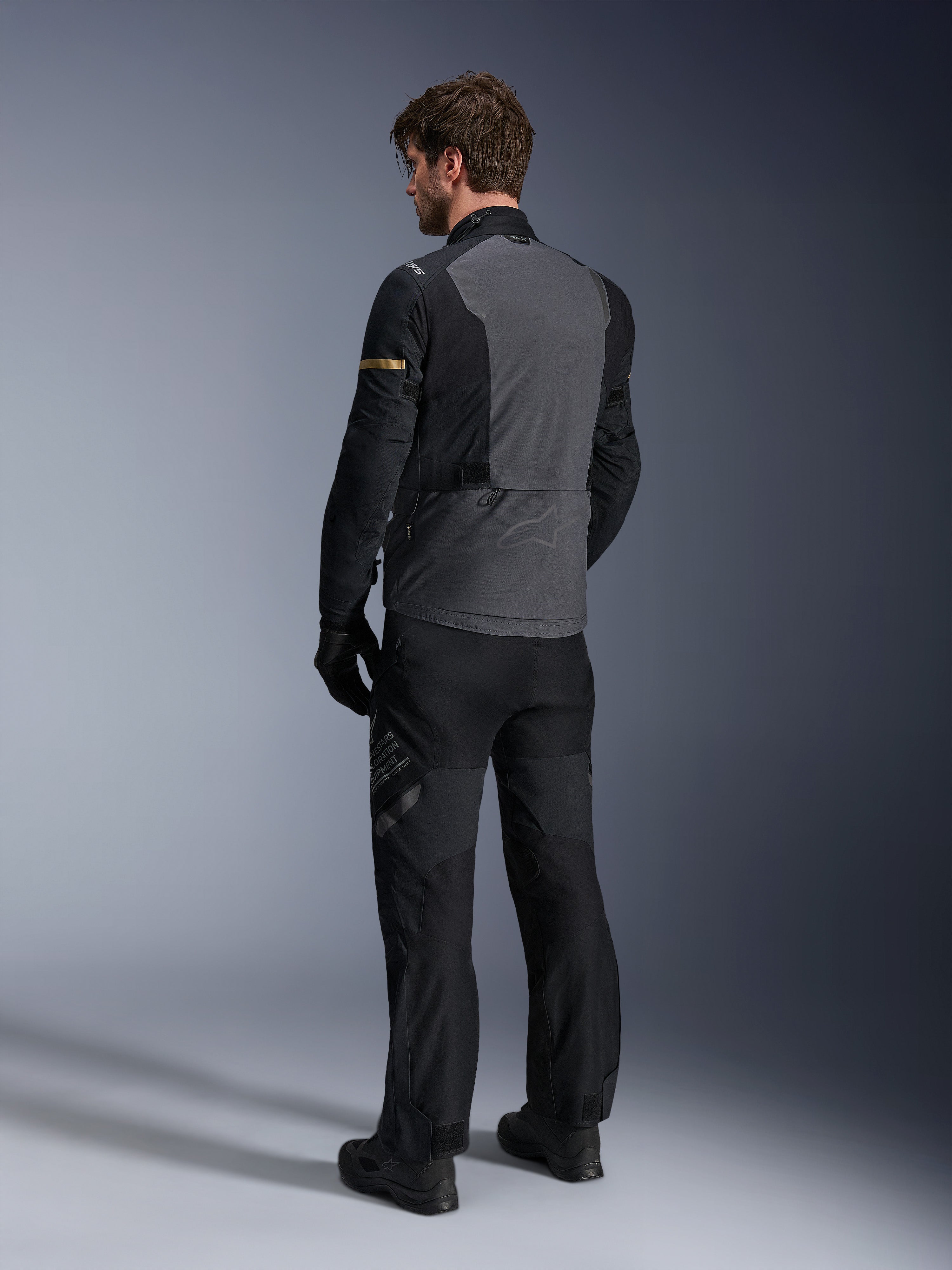 Alpinestars ST-7 2L GORE-TEX-jacka, mörkgrå och svart med mörka kaki-detaljer, vy bakifrån på en förare som bär jackan och matchande motorcykelbyxor, adventure touring-utrustning med vattentät laminerad konstruktion