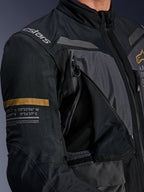 Alpinestars ST-7 2L GORE-TEX-jacka, mörkgrå och svart, närbild som visar en öppen ventilationspanel med dragkedja och mesh på bröstet, Alpinestars-logotyp på axeln och en kakifärgad rand på ärmen med tryckta geografiska koordinater, teknisk motorcykelutrustning för adventure-touring