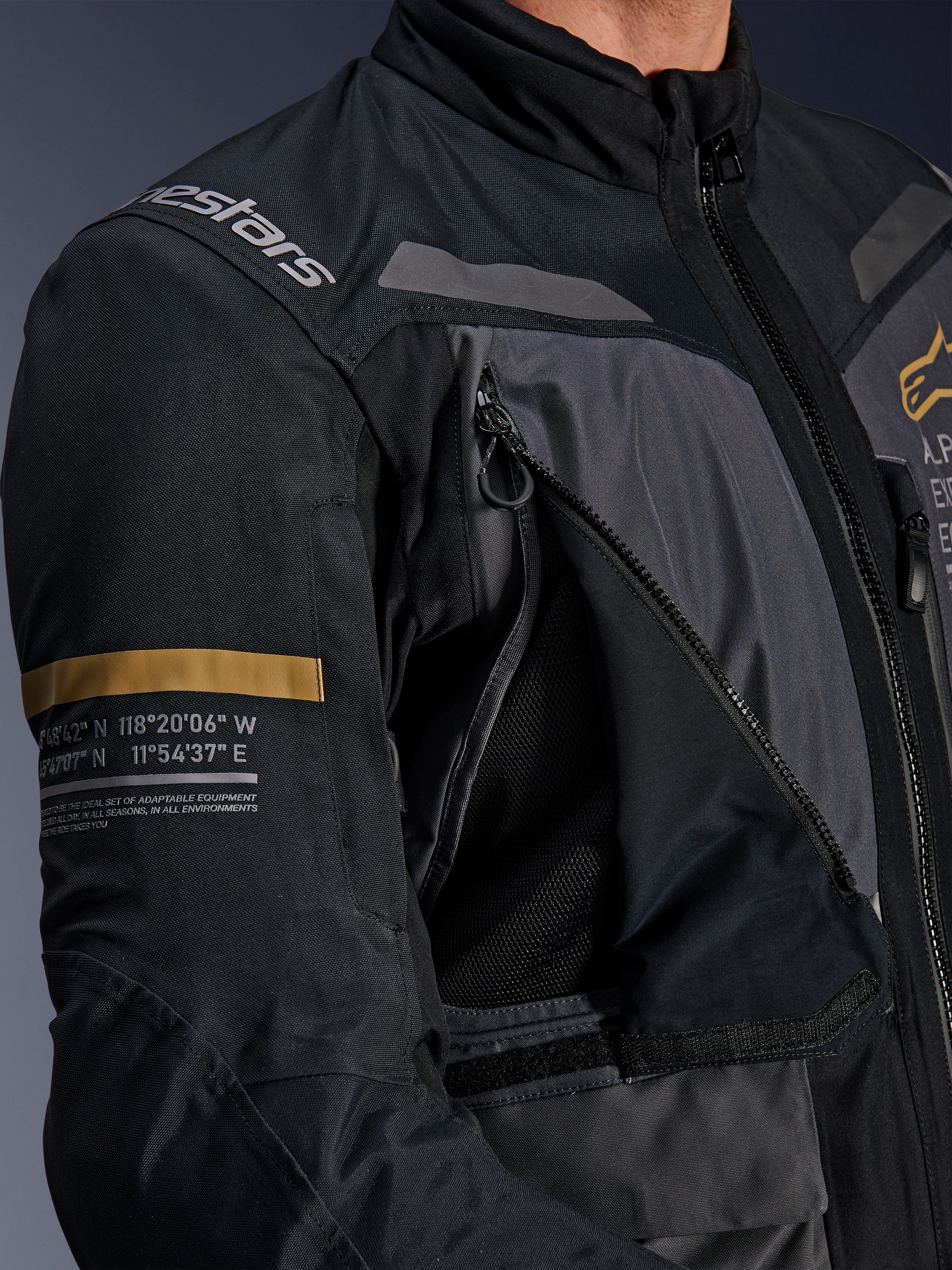 Alpinestars ST-7 2L GORE-TEX-jacka, mörkgrå och svart, närbild som visar en öppen ventilationspanel med dragkedja och mesh på bröstet, Alpinestars-logotyp på axeln och en kakifärgad rand på ärmen med tryckta geografiska koordinater, teknisk motorcykelutrustning för adventure-touring