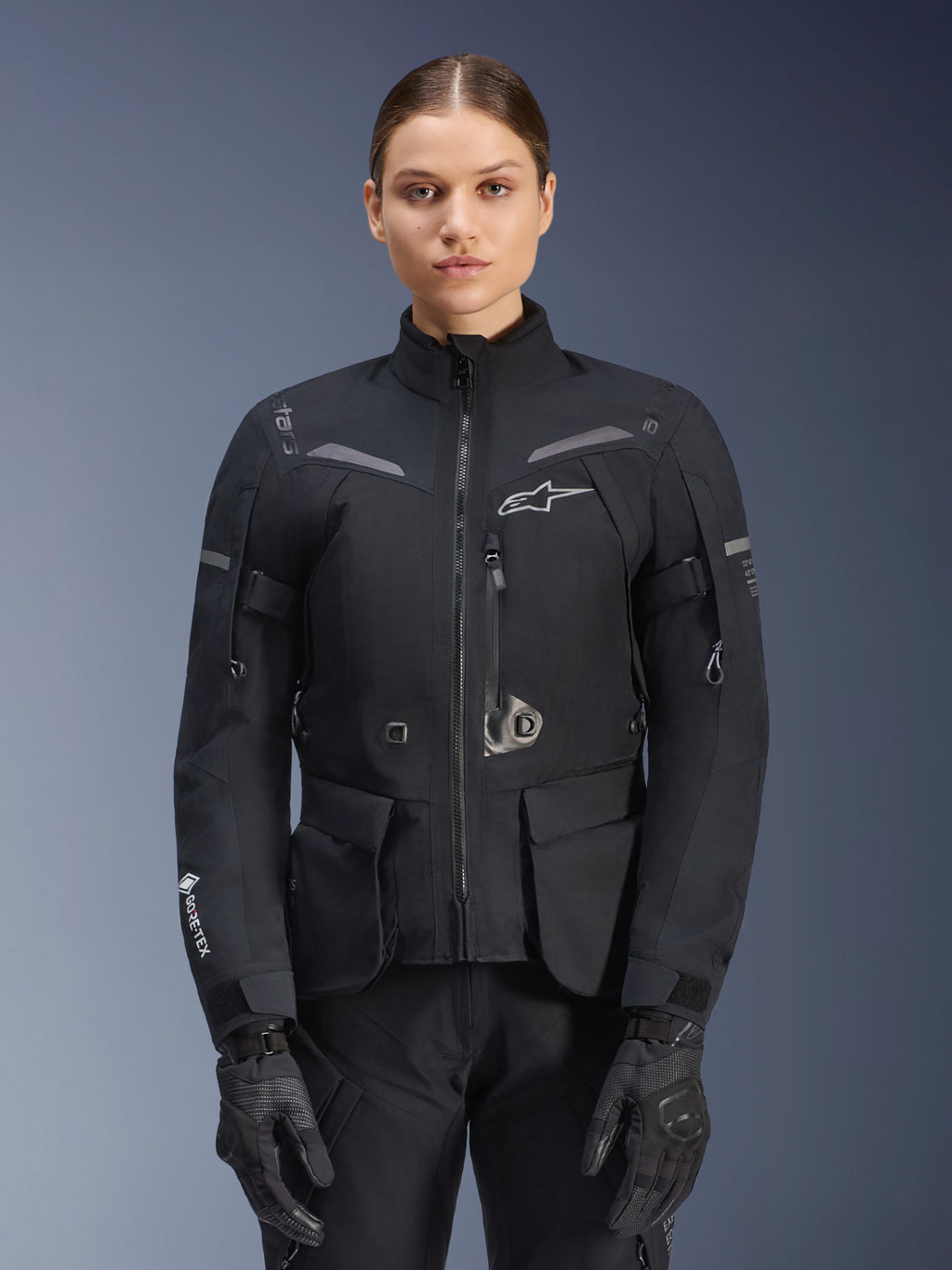 Kvinna klädd i Alpinestars Stella ST-7 2L GORE-TEX-jacka, svart med mörkgrå detaljer, touringjacka för motorcykel med stora cargofickor och hög krage, kombinerad med svarta handskar och byxor, vattentät MC-utrustning för äventyrskörning