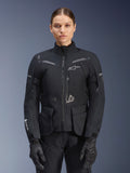 Kvinna klädd i Alpinestars Stella ST-7 2L GORE-TEX-jacka, svart med mörkgrå detaljer, touringjacka för motorcykel med stora cargofickor och hög krage, kombinerad med svarta handskar och byxor, vattentät MC-utrustning för äventyrskörning