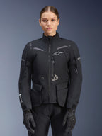 Kvinna klädd i Alpinestars Stella ST-7 2L GORE-TEX-jacka, svart med mörkgrå detaljer, touringjacka för motorcykel med stora cargofickor och hög krage, kombinerad med svarta handskar och byxor, vattentät MC-utrustning för äventyrskörning