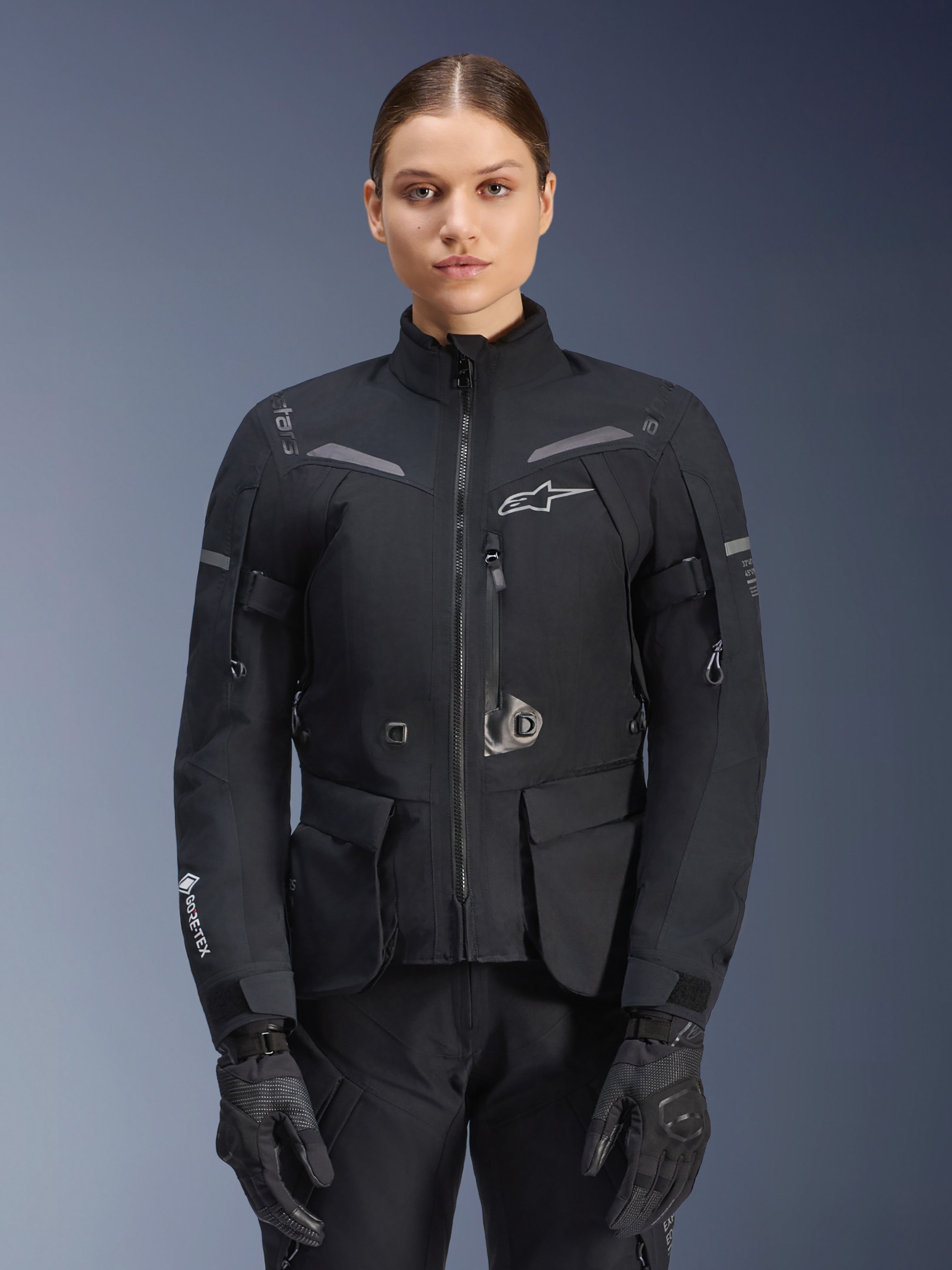 Kvinna klädd i Alpinestars Stella ST-7 2L GORE-TEX-jacka, svart med mörkgrå detaljer, touringjacka för motorcykel med stora cargofickor och hög krage, kombinerad med svarta handskar och byxor, vattentät MC-utrustning för äventyrskörning