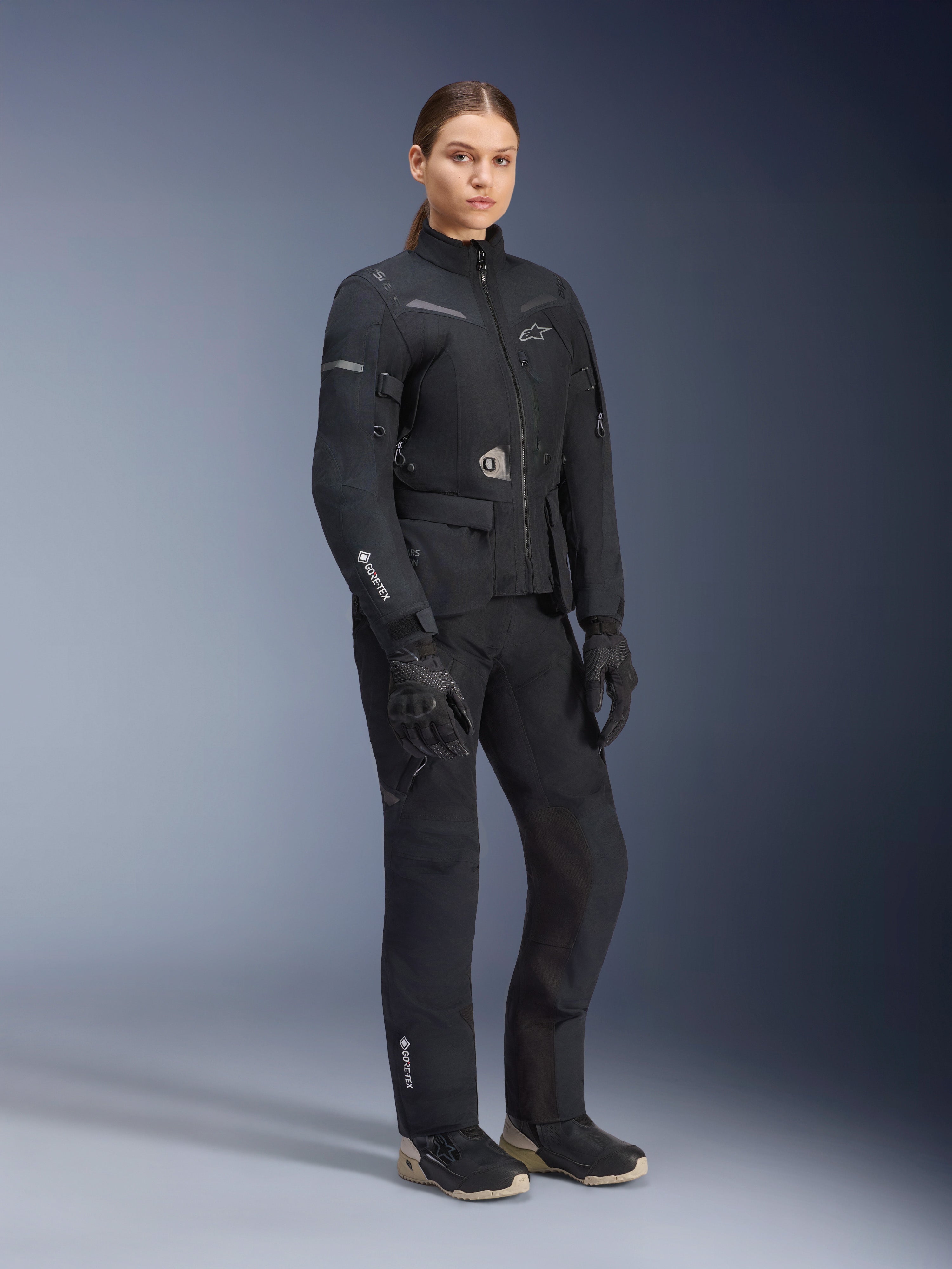 Kvinnlig modell bär Alpinestars Stella ST-7 2L GORE-TEX adventure touring-ställ, svart och mörkgrå jacka och byxor, med tydlig GORE-TEX-branding på ärmen och benet, kombinerat med svarta handskar och MC-stövlar, professionell motorcykelutrustning designad för touring i alla väder.