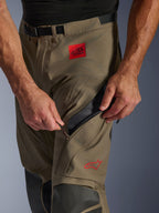 Alpinestars Nazca 3L GORE-TEX PRO-byxor i Walnut Black, närbild på lårområdet som visar en hand som öppnar det stora ventilationsintaget med dragkedja, med ett rött ALL TERRA-märke och röd Alpinestars-logotyp, teknisk MC-utrustning för äventyrskörning.