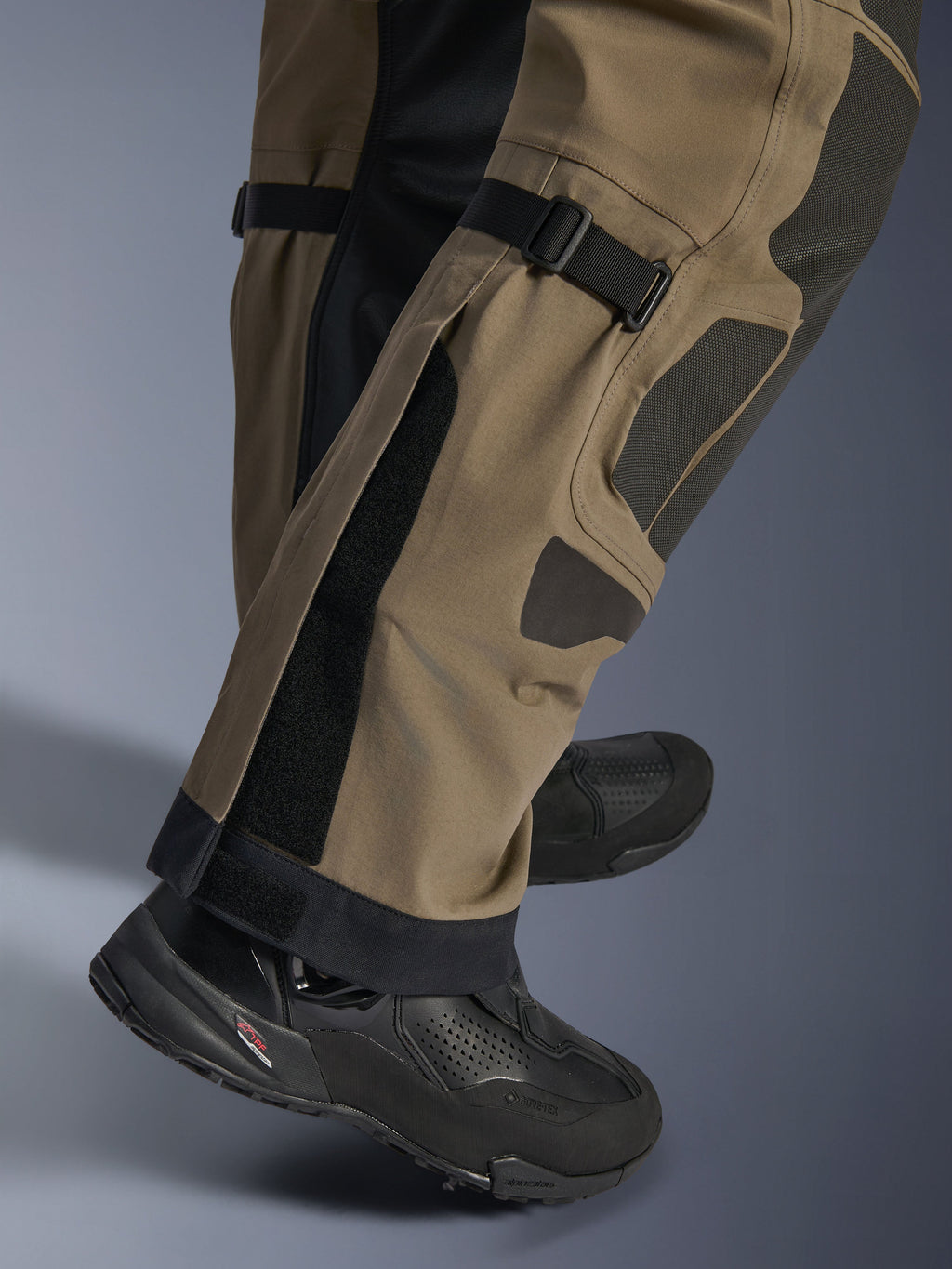 Närbild på Alpinestars Nazca 3L GORE-TEX PRO-byxor i Walnut Black, visar tekniska detaljer på underbenet inklusive justerare för vadvolym och Superfabric-förstärkningar, bärs med svarta äventyrsmotorcykelstövlar.
