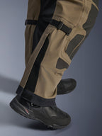 Närbild på Alpinestars Nazca 3L GORE-TEX PRO-byxor i Walnut Black, visar tekniska detaljer på underbenet inklusive justerare för vadvolym och Superfabric-förstärkningar, bärs med svarta äventyrsmotorcykelstövlar.