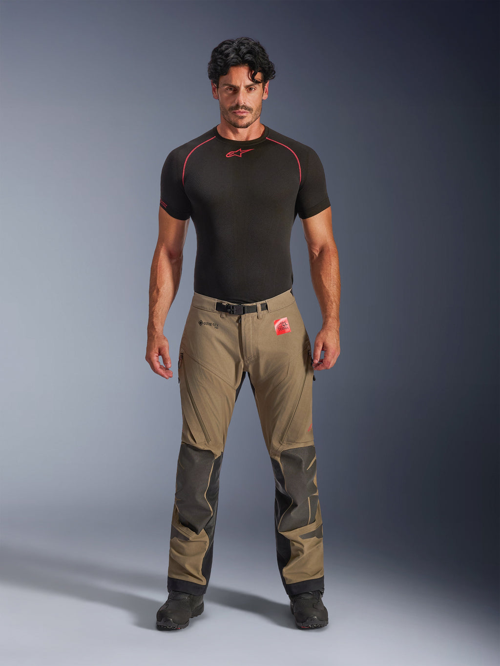 Manlig modell bär Alpinestars Nazca 3L GORE-TEX PRO-byxor i valnötsbrunt och svart, teknisk äventyrsutrustning med Superfabric-knäförstärkningar och ett magnetiskt midjespänne, kombinerat med en svart Alpinestars-undertröja, studiovy framifrån.
