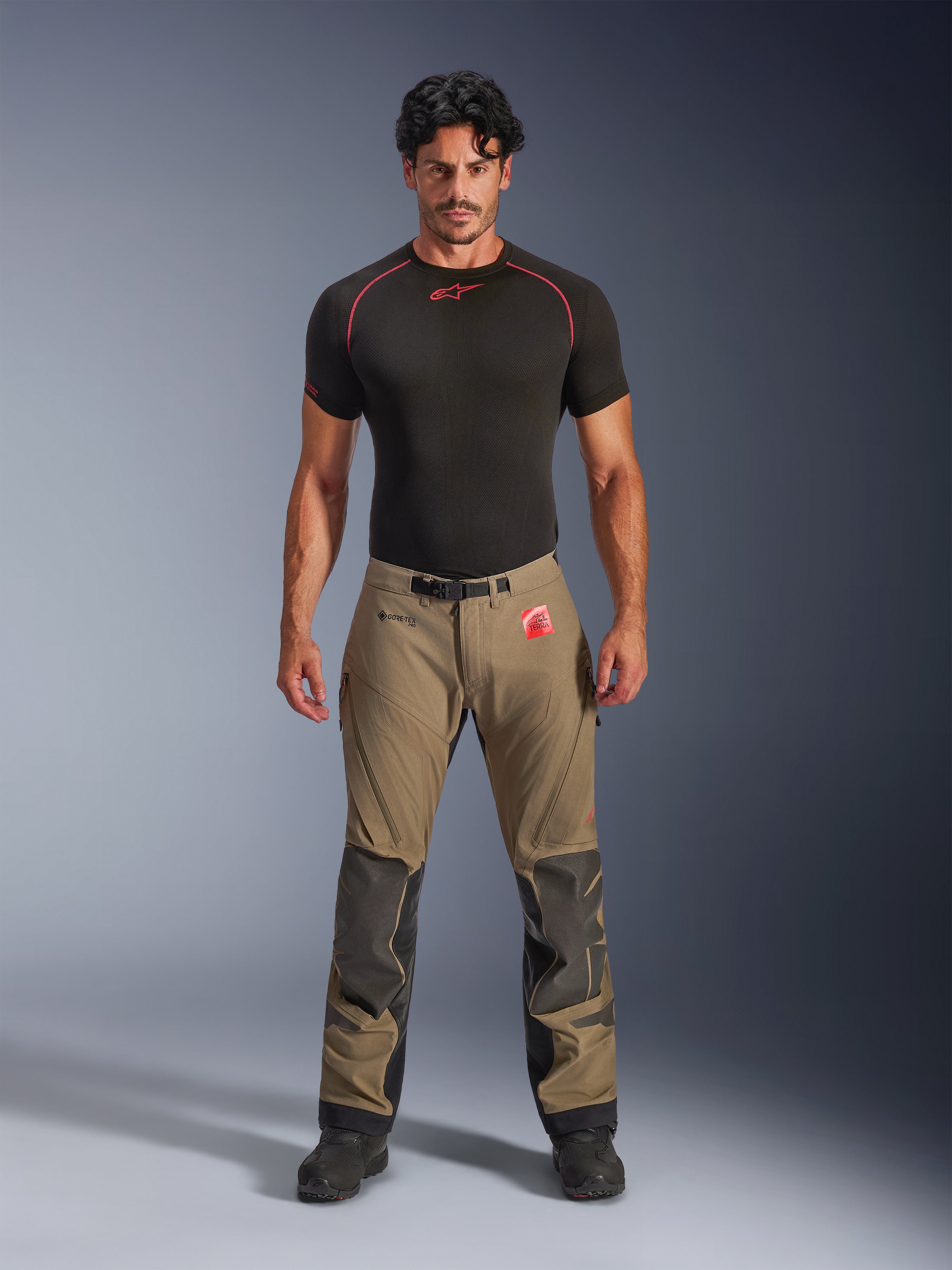 Manlig modell bär Alpinestars Nazca 3L GORE-TEX PRO-byxor i valnötsbrunt och svart, teknisk äventyrsutrustning med Superfabric-knäförstärkningar och ett magnetiskt midjespänne, kombinerat med en svart Alpinestars-undertröja, studiovy framifrån.