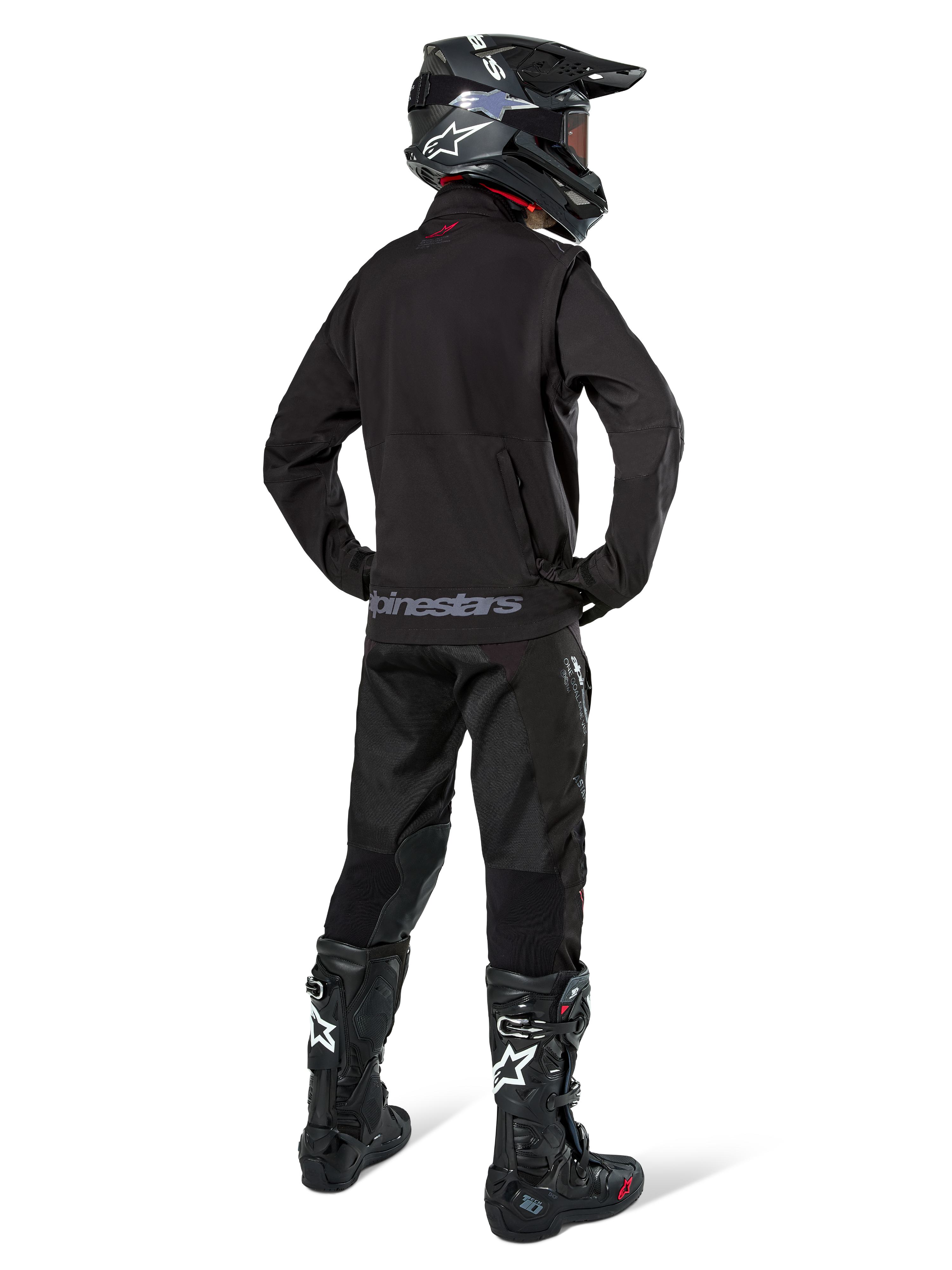 Enduro rider viewed from the back wearing the Alpinestars Lite-Dura Softshell Jacket in Black, paired with black technical riding pants and boots, featuring a zippered rear storage pocket and Alpinestars logo on the lower hem, complete off-road gear for motocross.
