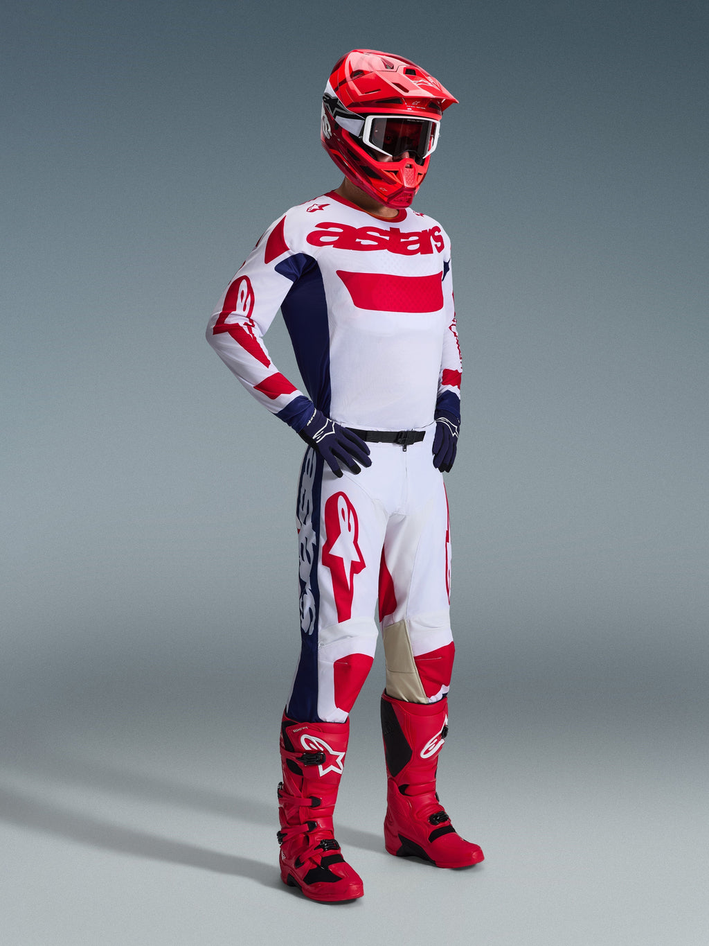 Motocrossförare i komplett racingutrustning, Alpinestars 2026 Racer Riway-byxor i vitt, blått och rött, med en vit bas, framträdande röda logotyper och blå sidopaneler, matchat med en vit och röd tröja, röd hjälm och röda motocross-stövlar.
