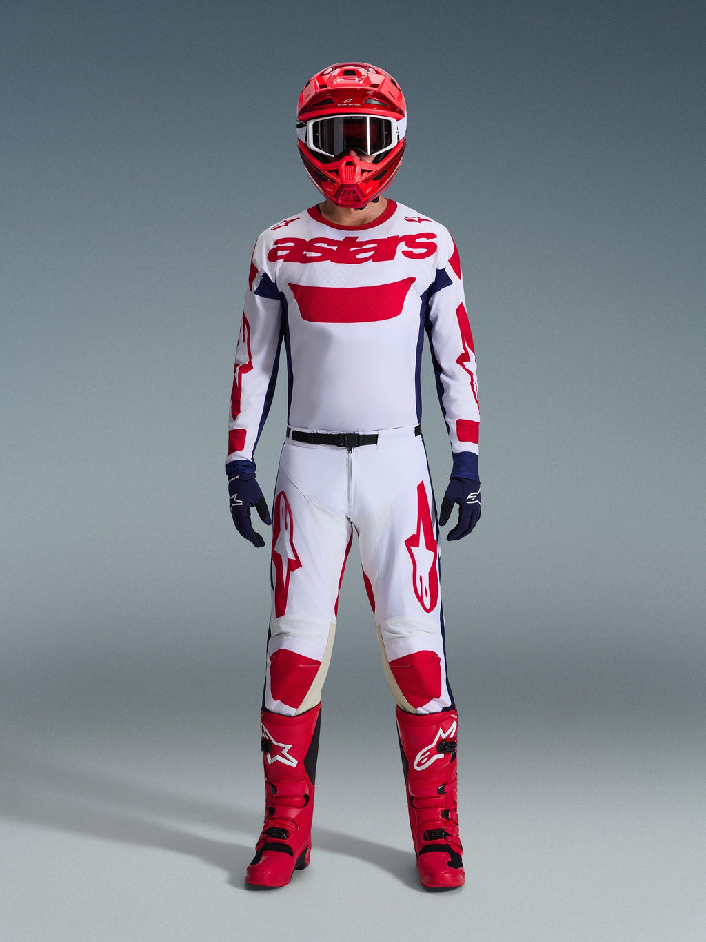 Motocrossförare i komplett racingutrustning, vit tröja med röd Alpinestars-branding och blå detaljer, Alpinestars 2026 Racer Riway-byxor i White Blue Red, röd hjälm med matchande goggles, röda och svarta stövlar, mörkblå handskar, koordinerad outfit för terrängkörning