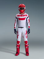 Motocrossförare i komplett racingutrustning, vit tröja med röd Alpinestars-branding och blå detaljer, Alpinestars 2026 Racer Riway-byxor i White Blue Red, röd hjälm med matchande goggles, röda och svarta stövlar, mörkblå handskar, koordinerad outfit för terrängkörning