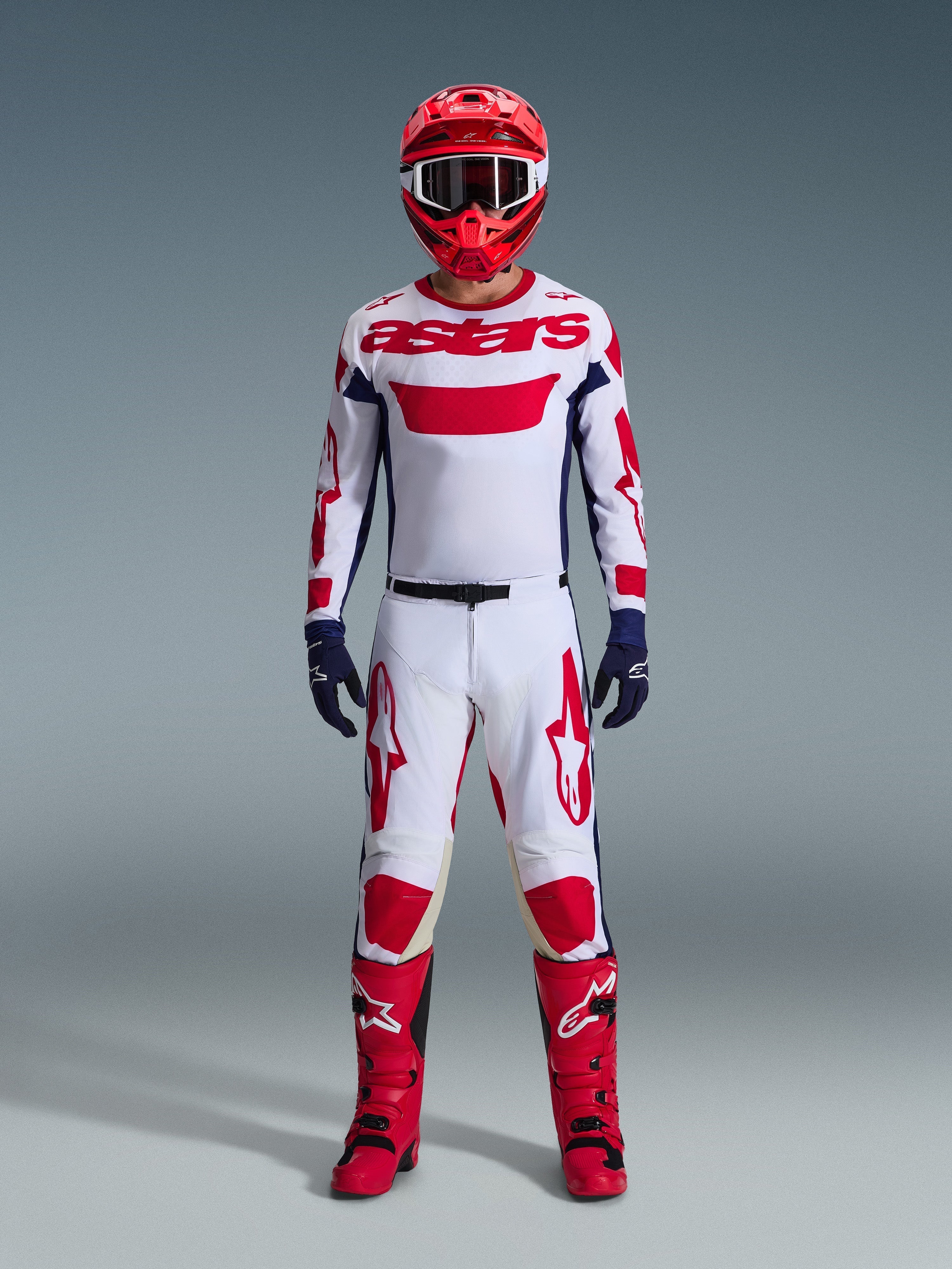 Motocrossförare i Alpinestars 2026 Racer Riway utrustningsset, vit tröja med röd "astars"-logotyp och blå sidopaneler, vita byxor med röd grafik, röd racinghjälm och stövlar, mörkblå handskar, komplett koordinerad motocrossutrustning