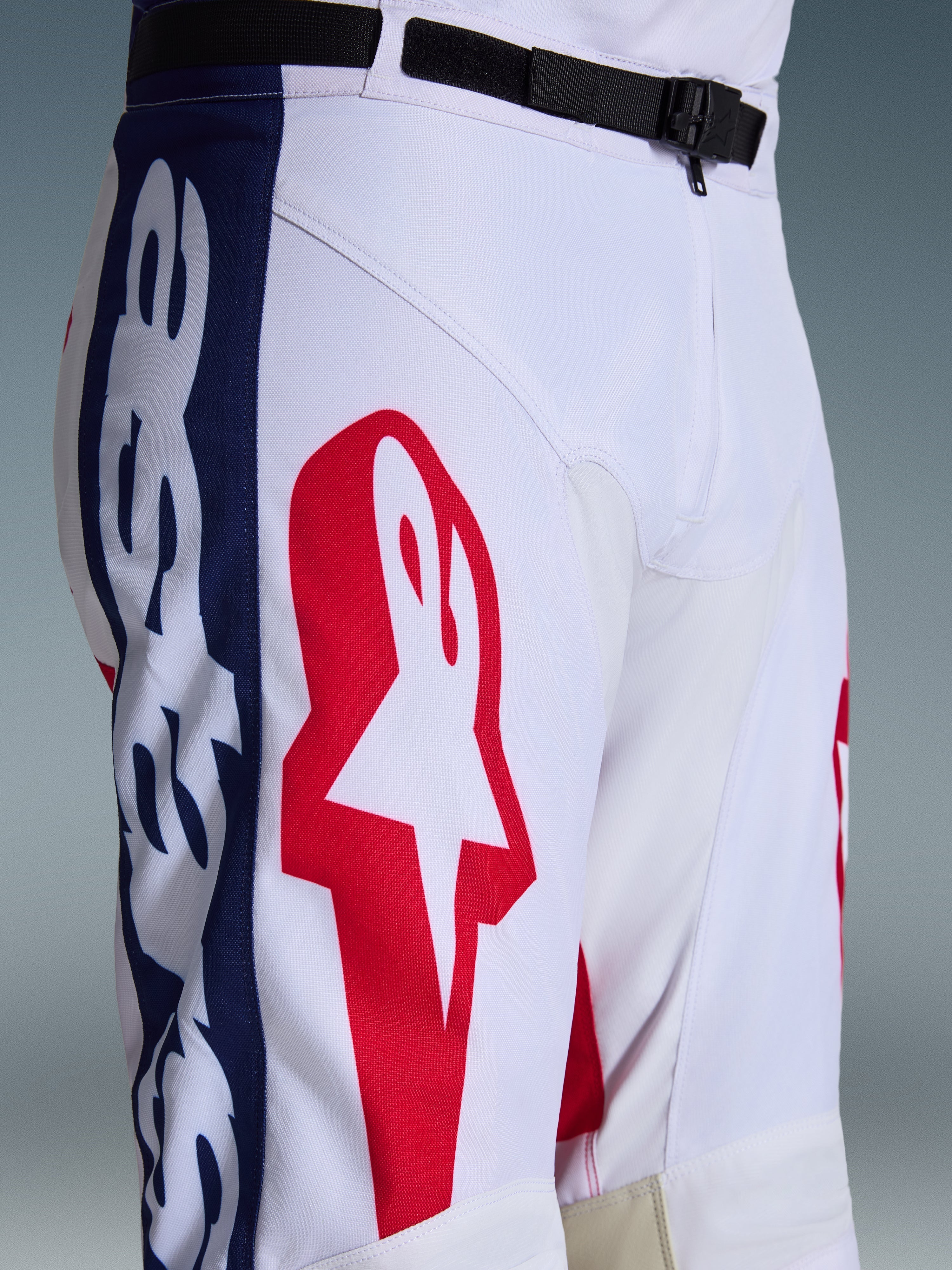 Alpinestars 2026 Racer Riway byxor, motocrossbyxor, vit, blå och röd, närbild av övre delen av benet och midjan som visar en stor röd stjärnlogotyp på vitt polytyg, blå sidopaneler med branding och svart justerbart midjebälte med SX2-magnetspänne, sportig racingdesign för terrängkörning.