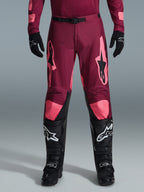 Alpinestars 2026 Racer Riway byxor, motocrossbyxor, mörkröd korall, bärs med svarta handskar och stövlar, med stora korallfärgade Alpinestars-logotyper på låren, svart midjespänne och förböjd benskärning för terrängkörning
