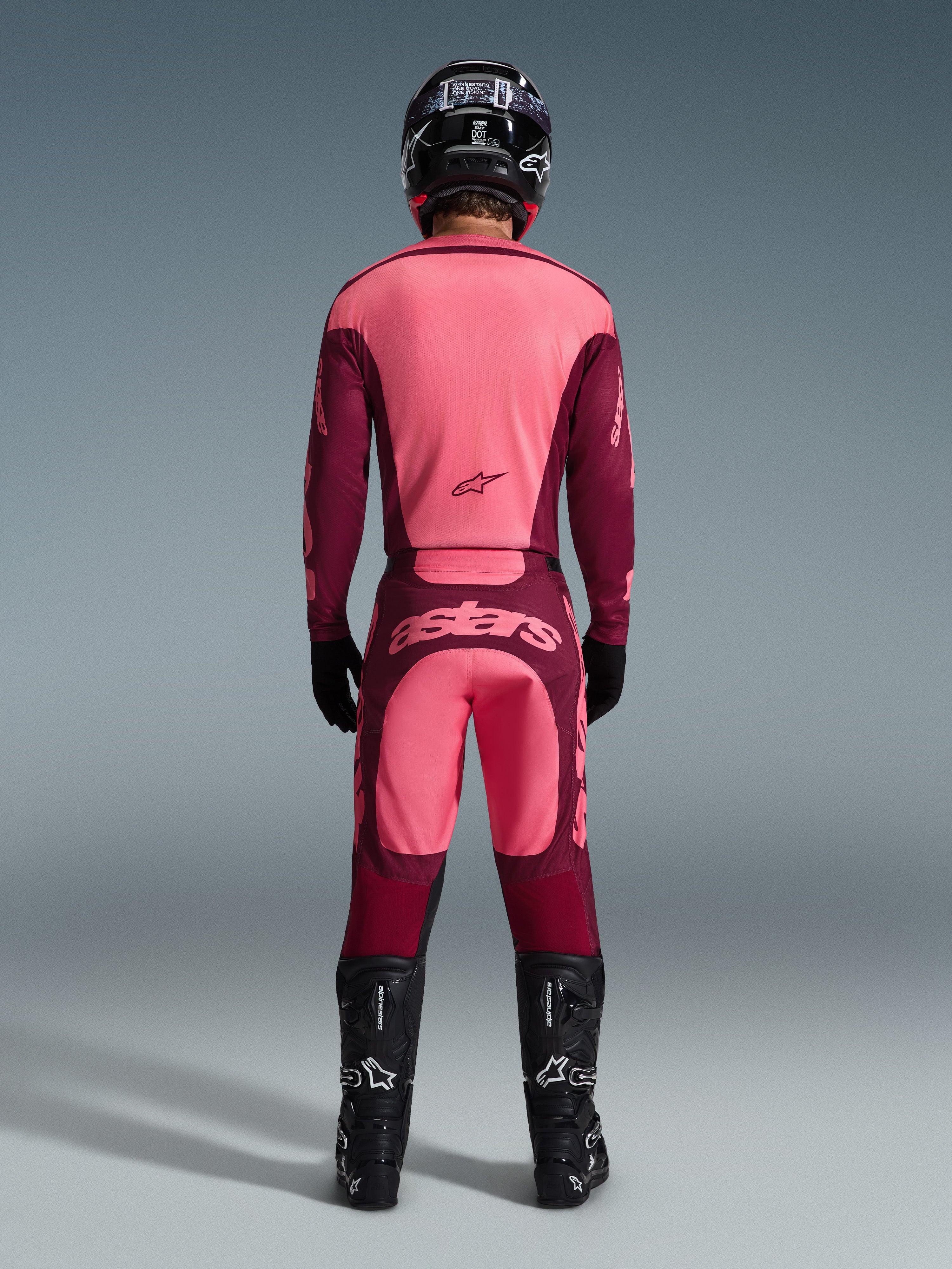 Motocrossförare sedd bakifrån iförd Alpinestars 2026 Racer Riway-byxor i Dark Red Coral, matchande tröja, svart hjälm, svarta handskar och svarta Alpinestars-stövlar, med en framträdande 'astars'-logotyp på sätet och en tvåtonad röd design.