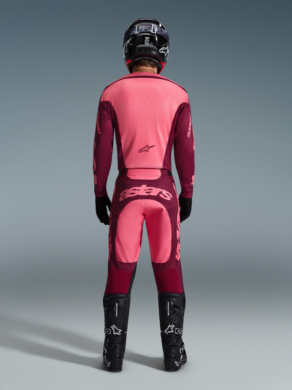 Motocrossförare sedd bakifrån klädd i Alpinestars 2026 Racer Riway-tröja och matchande byxor i Dark Red Coral, med svarta stövlar, en svart hjälm och svarta handskar, koordinerad högpresterande utrustning för terrängkörning