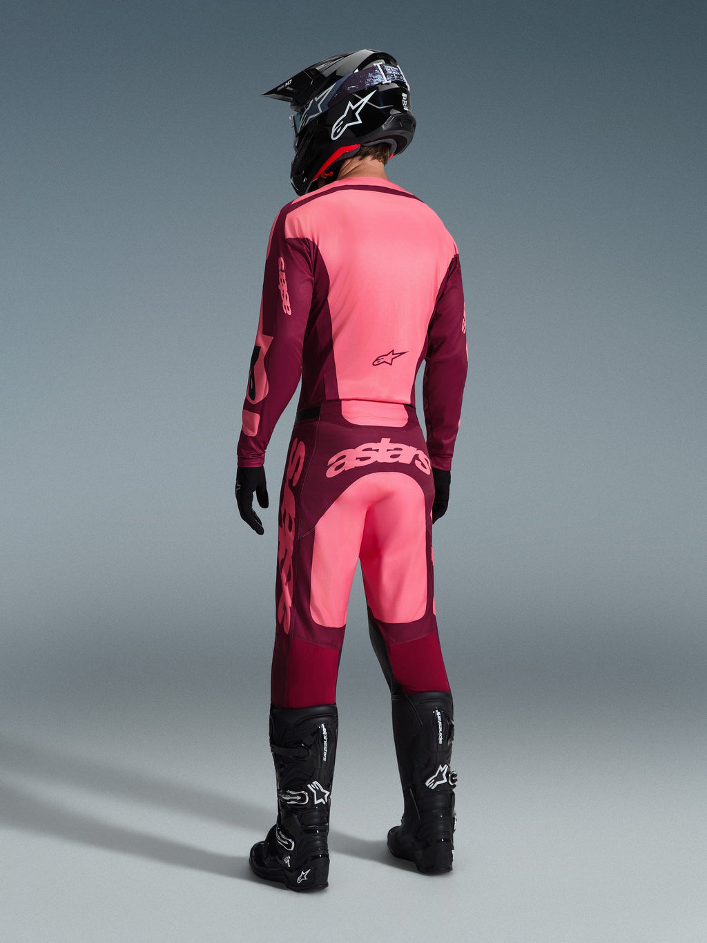 Alpinestars 2026 Racer Riway-byxor, motocrossbyxor, Dark Red Coral, vy bakifrån av en förare i matchande tröja, svart hjälm och svarta stövlar, med framträdande korallfärgad logotyp på sätet och ergonomisk atletisk passform för terrängkörning