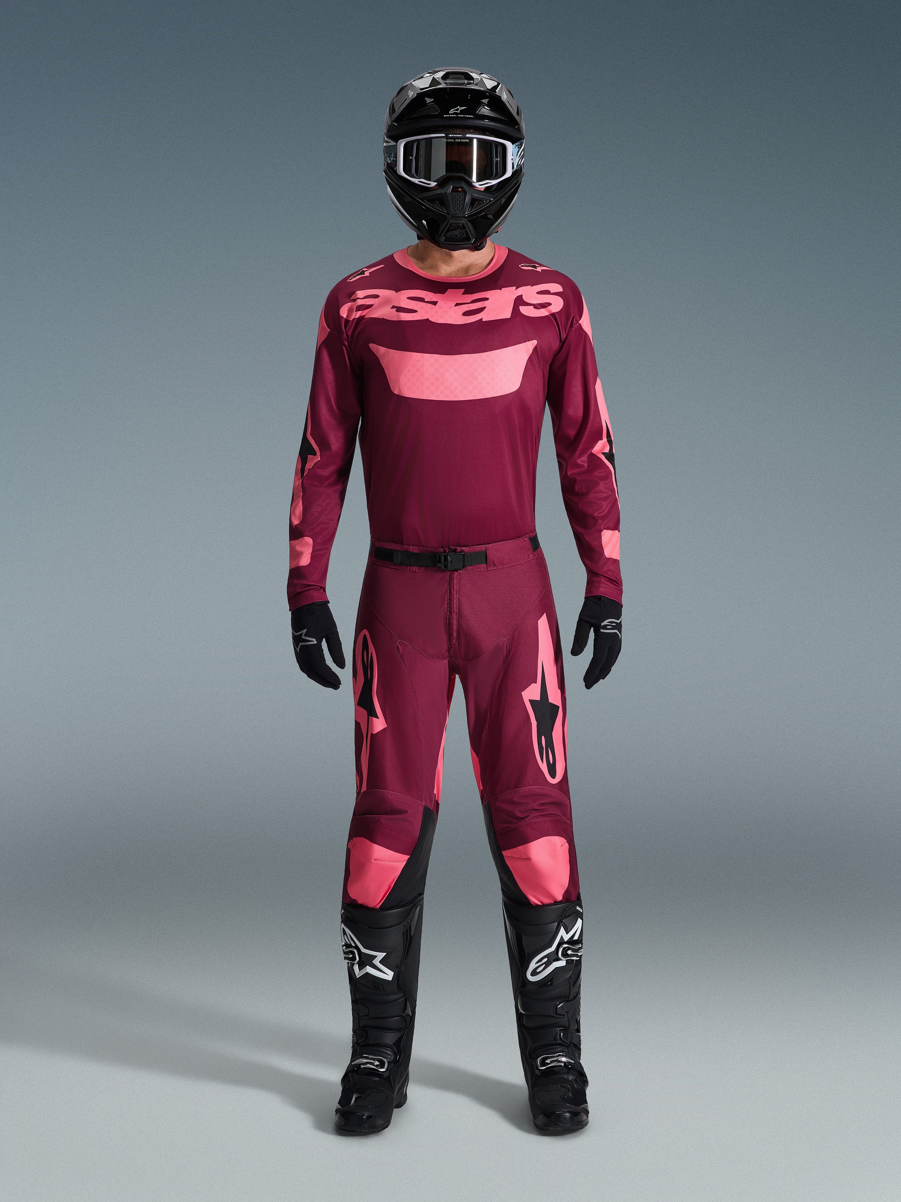 Motocrossförare klädd i Alpinestars 2026 Racer Riway-utrustningsset, vinröd tröja med rosa "astars"-logotyp, matchande vinröda byxor med rosa detaljer på knän och ben, svart hjälm, svarta handskar och svarta stövlar, komplett prestandautrustning för terrängkörning