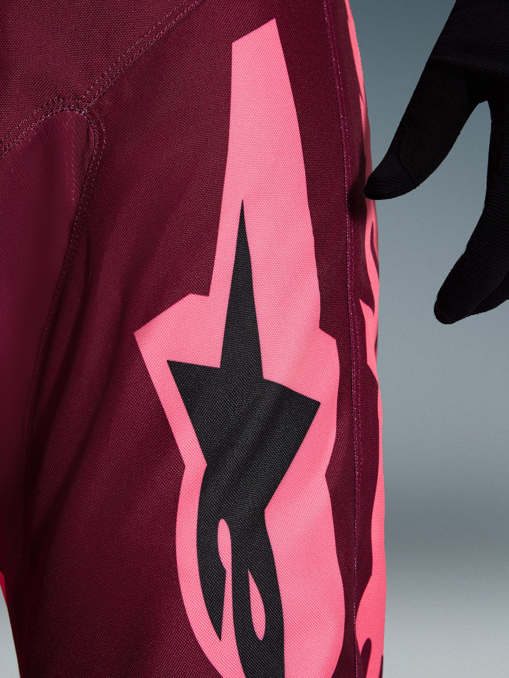 Alpinestars 2026 Racer Riway-byxor, motocrossbyxor, Dark Red Coral, närbild som visar en stor korallrosa vinkelformad grafik och svart stjärnlogotyp, som framhäver den slitstarka texturen i polytyg och prestandasömmar