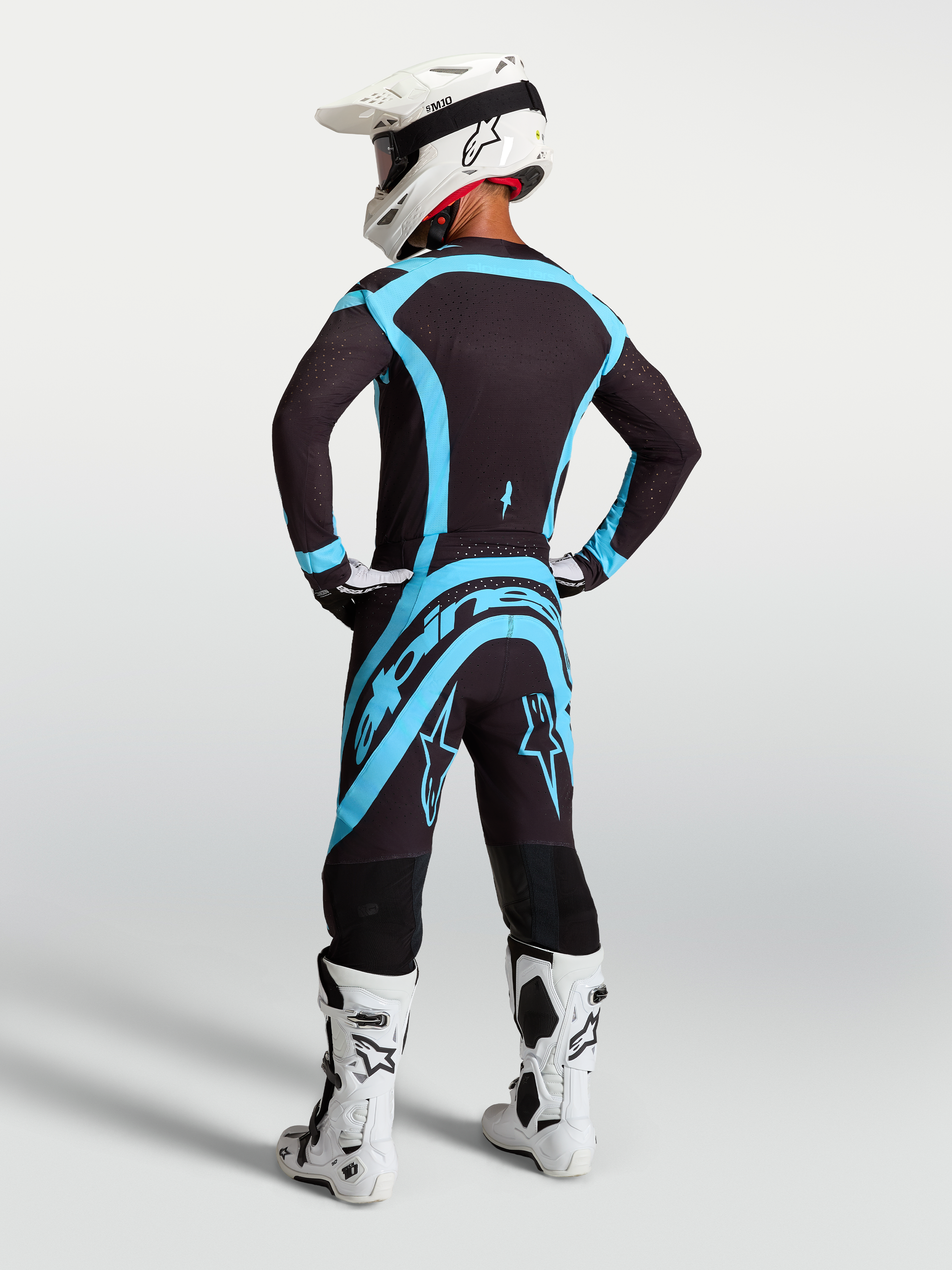 Alpinestars Supertech Vesta byxor, professionella motocrossbyxor i svart och turkos, vy bakifrån på modell med vit hjälm och vita stövlar, högpresterande utrustning för tävling och terrängkörning