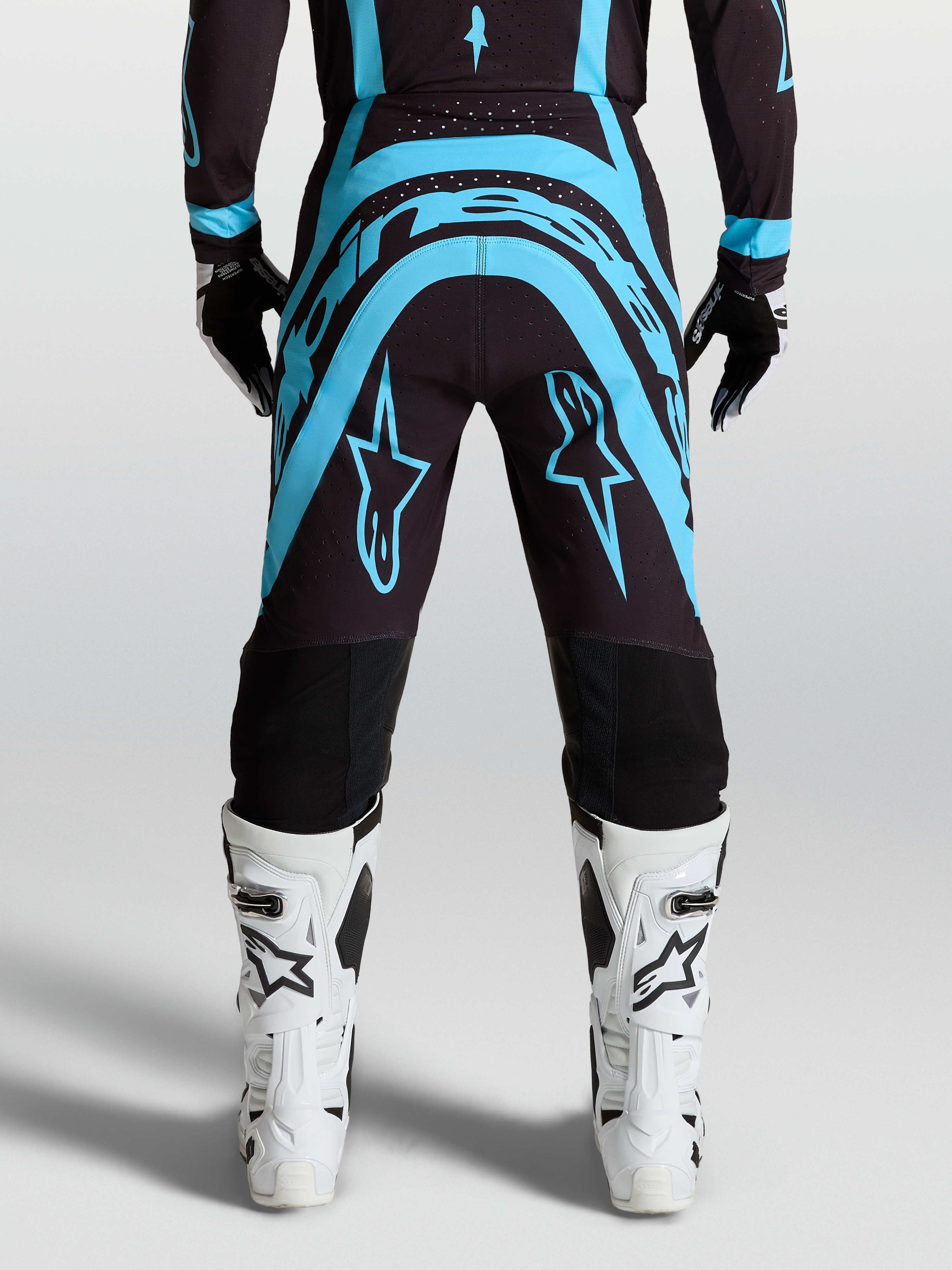 Alpinestars Supertech Vesta byxor, svarta och turkosa motocrossbyxor med vita motorcykelstövlar, vy bakifrån.