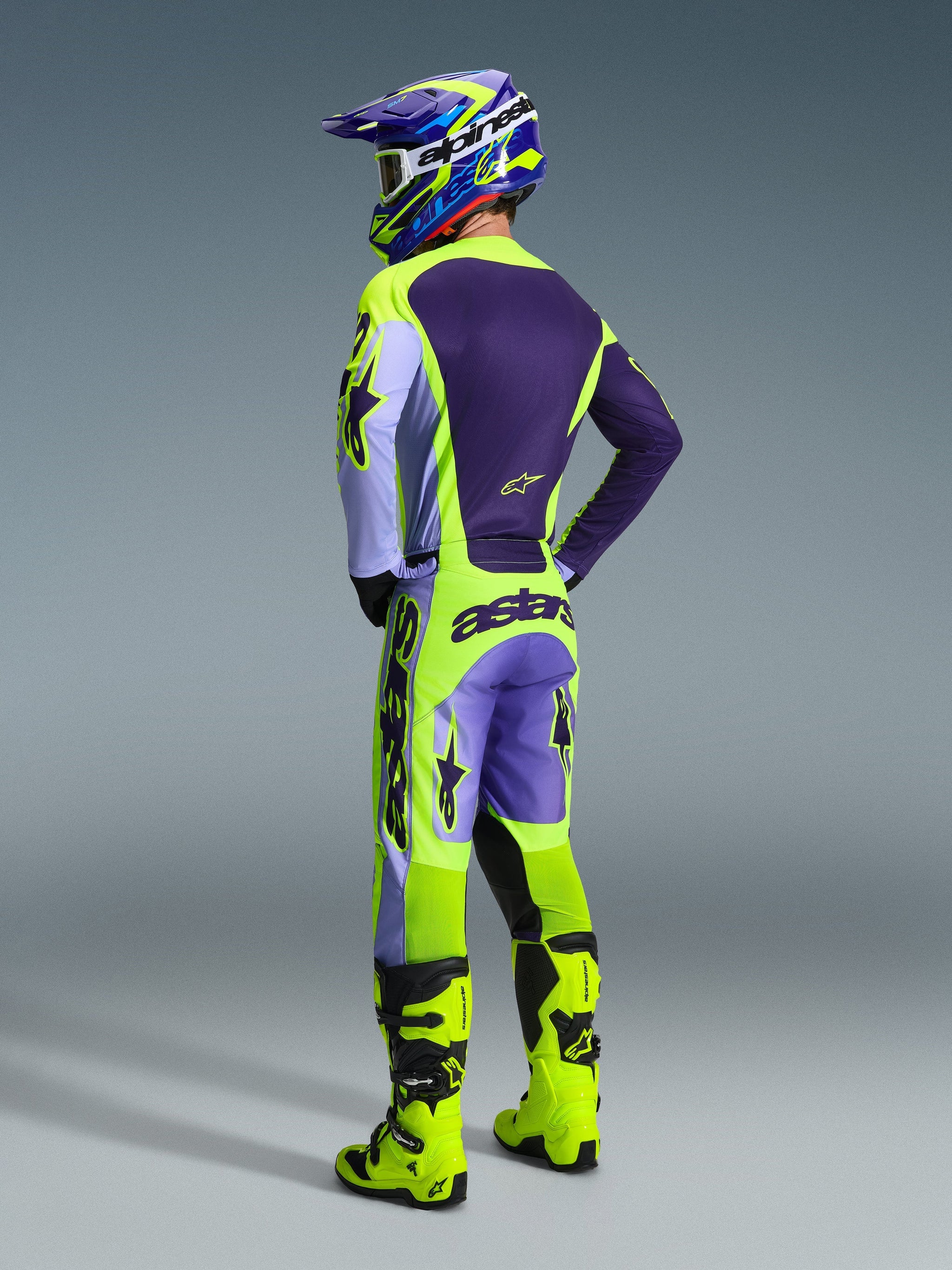 Motocrossförare sedd bakifrån iklädd Alpinestars 2026 Racer Portl-byxor i lila och neongult (Purple Yellow Fluo), matchad med en lila och gul tröja, hjälm och klargula racingstövlar, med tydlig 'astars'-logotyp på sätet och stjärnsymboler på benen.
