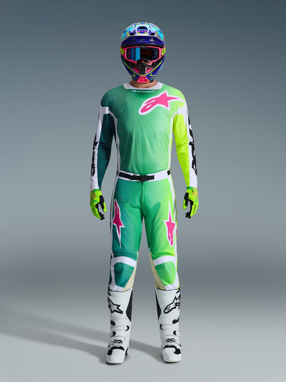Alpinestars 2026 Racer Portl Gear Set, motocross rider wearing complete racing gear, green jersey and pants with pink and white graphics, blue multi-colored helmet, white and black boots, and green gloves, coordinated outfit for off-road racing