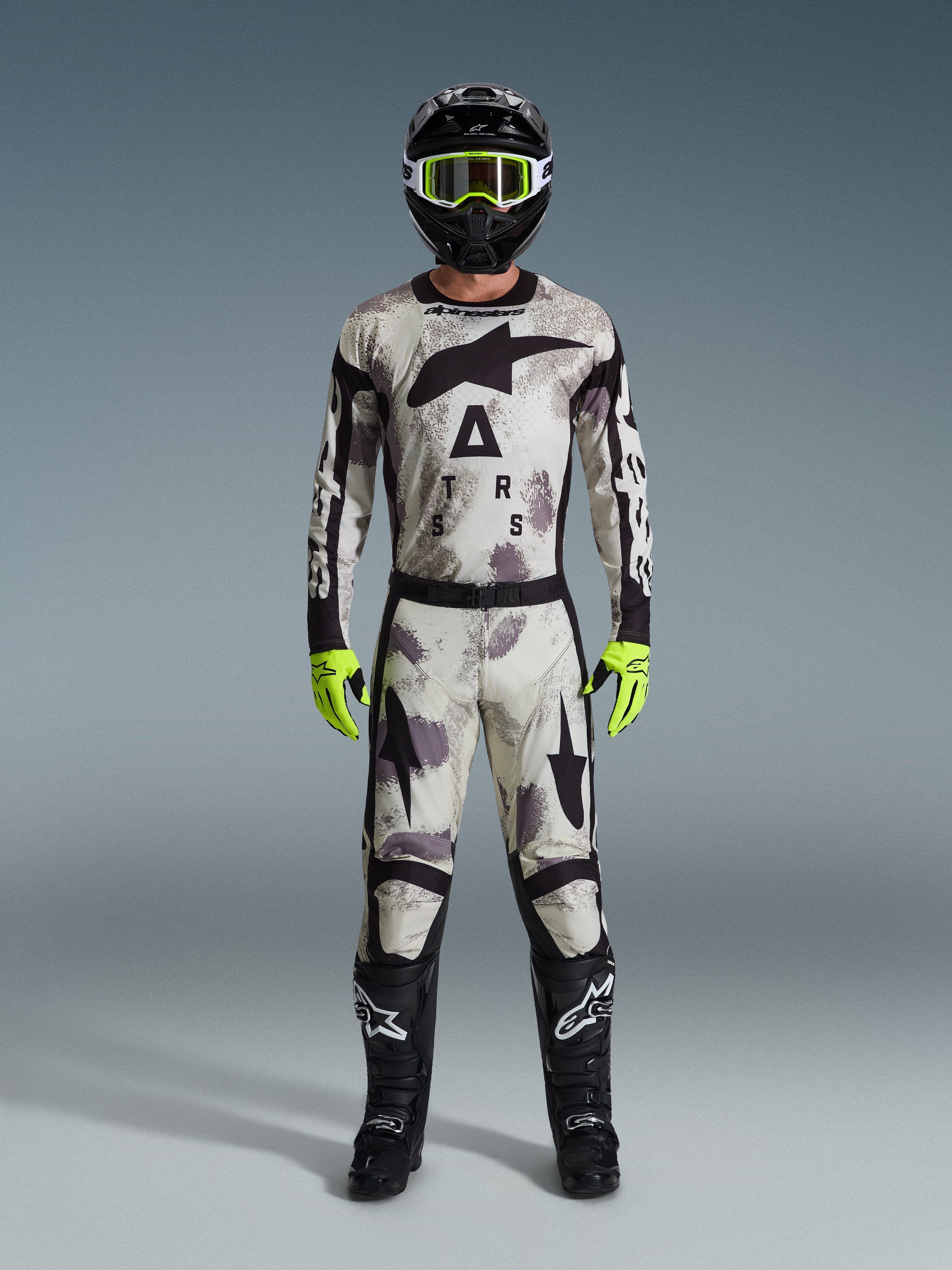 Motocross rider wearing the Alpinestars 2026 Racer Lahnd Gear Set, Sand Camo patterned suit with white, gray, and black design, black helmet with neon green goggles, bright neon green gloves, and black boots with white logos, complete coordinated outfit for off-road racing